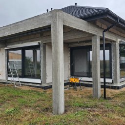 Adam Turowski OKNARO - Surowy stan budynku z zamontowanymi oknami i roletami zewnętrznymi. Widoczny szary beton, drabina i narzędzia. Budowa w toku, pochmurne niebo w tle.
