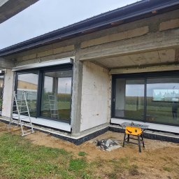 Adam Turowski OKNARO - Surowy stan budynku z zamontowanymi oknami z czarnymi ramami i roletami zewnętrznymi. Na zewnątrz drabina i stół roboczy z narzędziami.