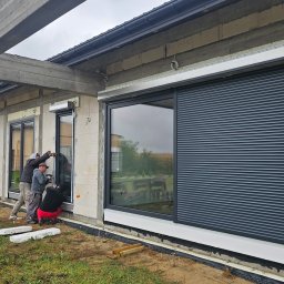 Adam Turowski OKNARO - Montaż okien i rolet zewnętrznych w surowym budynku. Widoczne okna z zamontowanymi roletami, drabina i trzech monterów podczas pracy. Budynek w trakcie budowy.