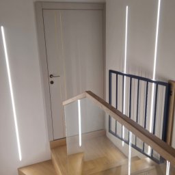 Adam Turowski OKNARO - Nowoczesne schody drewniane z balustradą ze szkła i drewna, oświetlone pionowymi lampami LED na ścianie. Widoczny fragment drzwi wewnętrznych z delikatnymi zdobieniami.