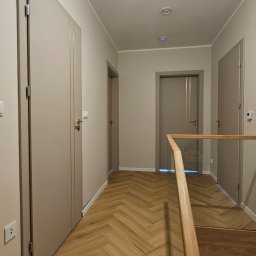 Adam Turowski OKNARO - Korytarz z parkietem w jodełkę i beżowymi drzwiami. Widoczne trzy pary drzwi, oświetlenie sufitowe i drewniana balustrada. Minimalistyczny design.