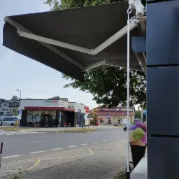 Zewnętrzna markiza rozkładana nad wejściem do lokalu, widok na ulicę z budynkami i drzewami w tle.