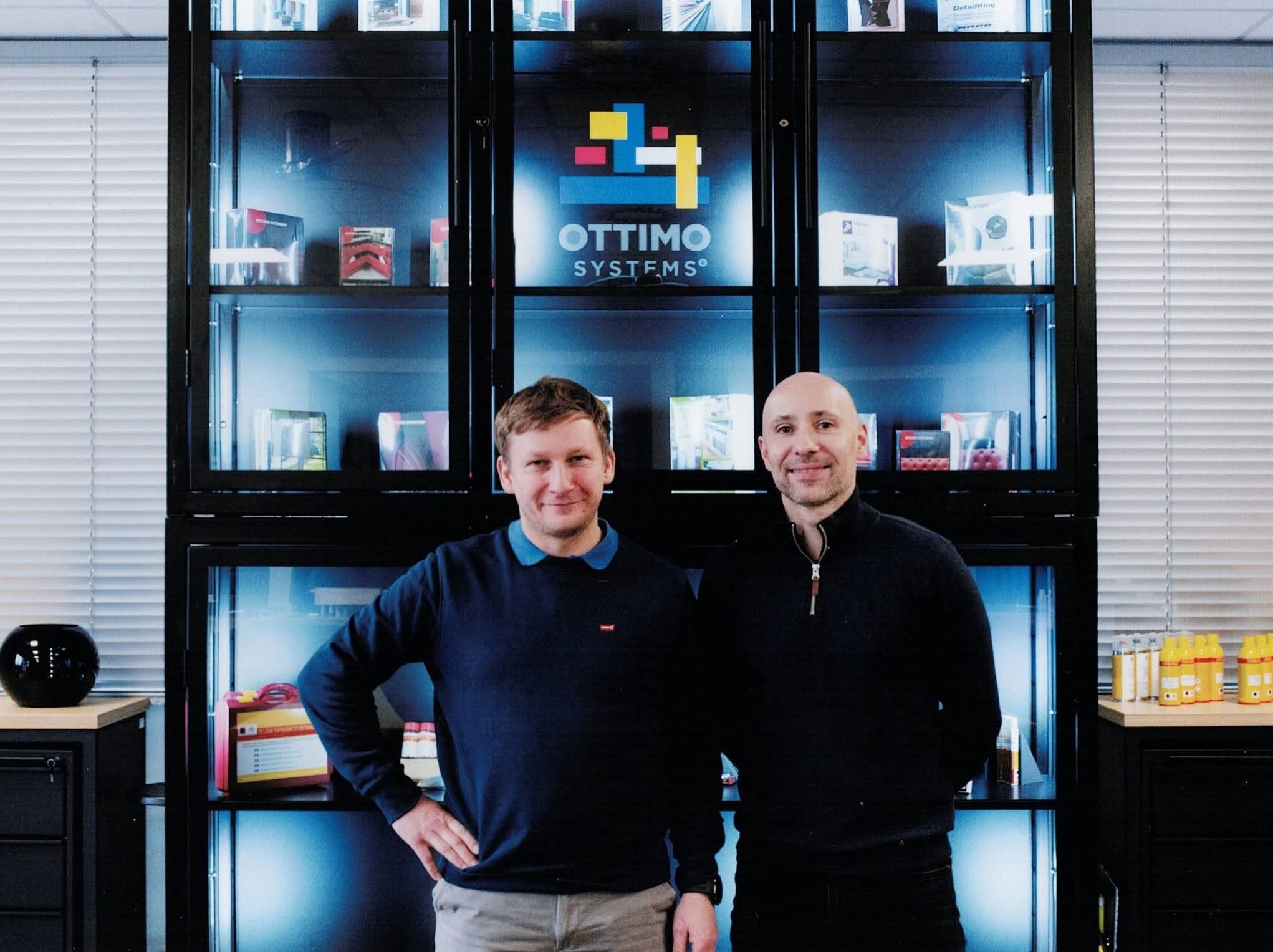 Dwóch mężczyzn na tle regału z produktami Ottimo Systems, oświetlonego niebieskim światłem, z żaluzjami w tle.