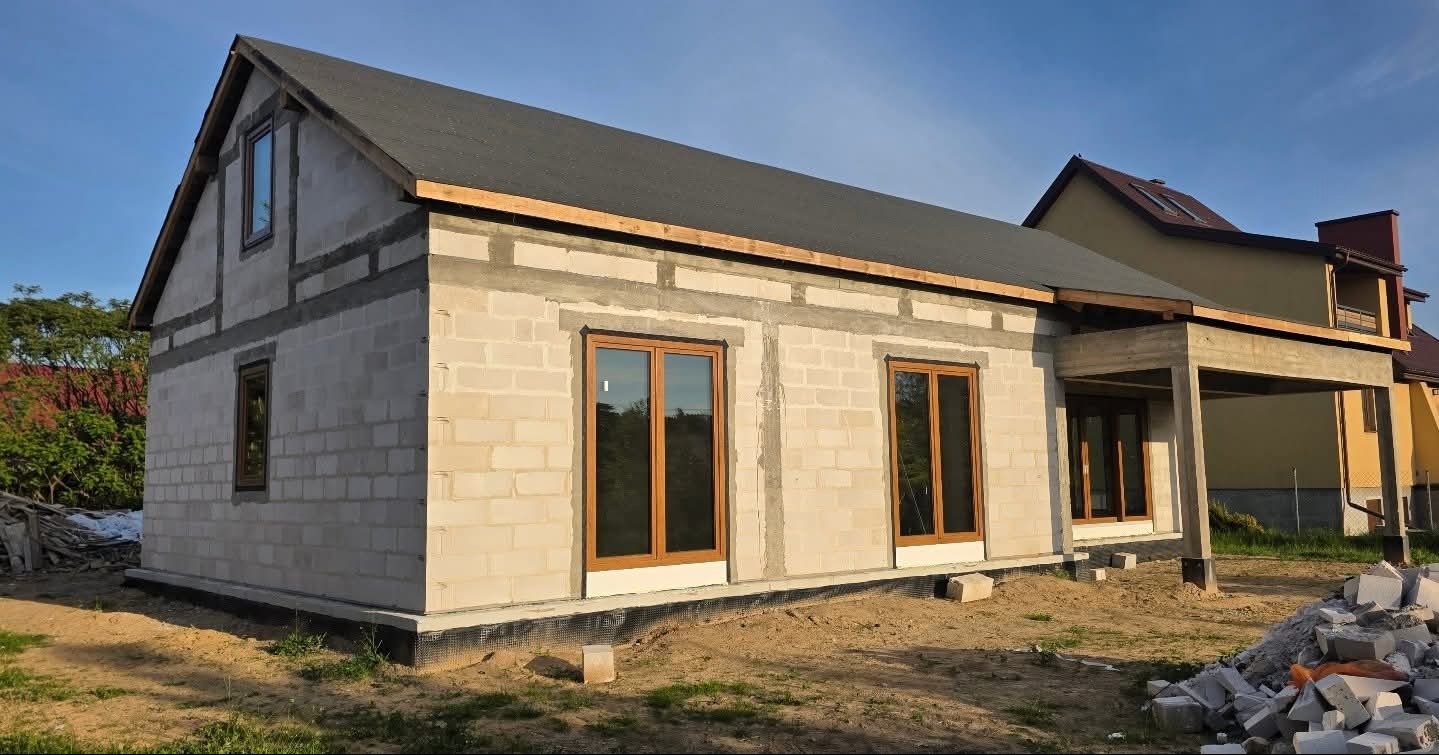 Surowy stan domu jednorodzinnego z zamontowanymi oknami w brązowych ramach, widoczne bloczki betonowe i przygotowane podłoże pod elewację.