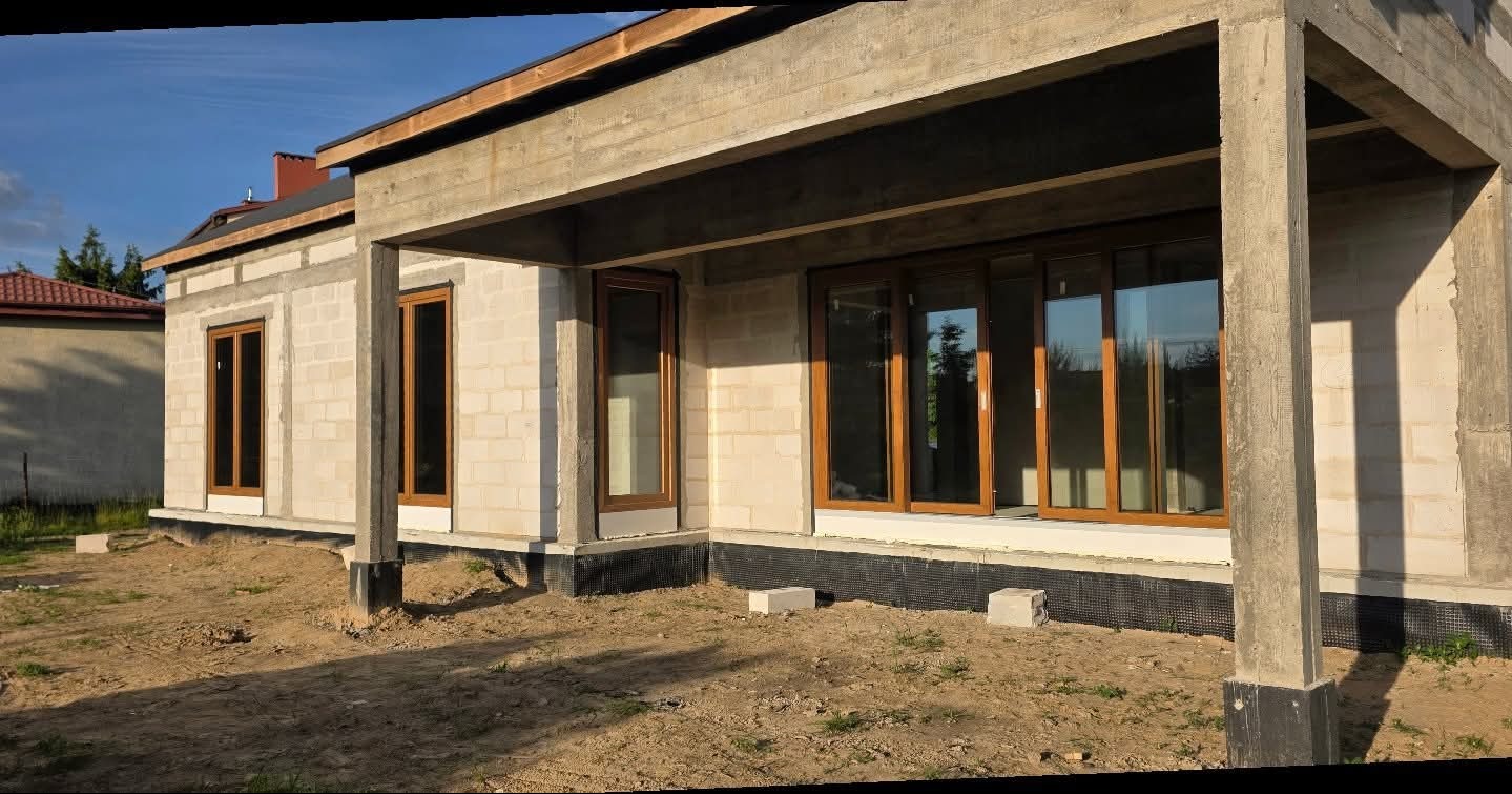 Surowy stan domu z zamontowanymi oknami z brązowymi ramami. Widoczne bloczki betonowe i betonowe filary. Dom w trakcie budowy, teren wokół niezagospodarowany.