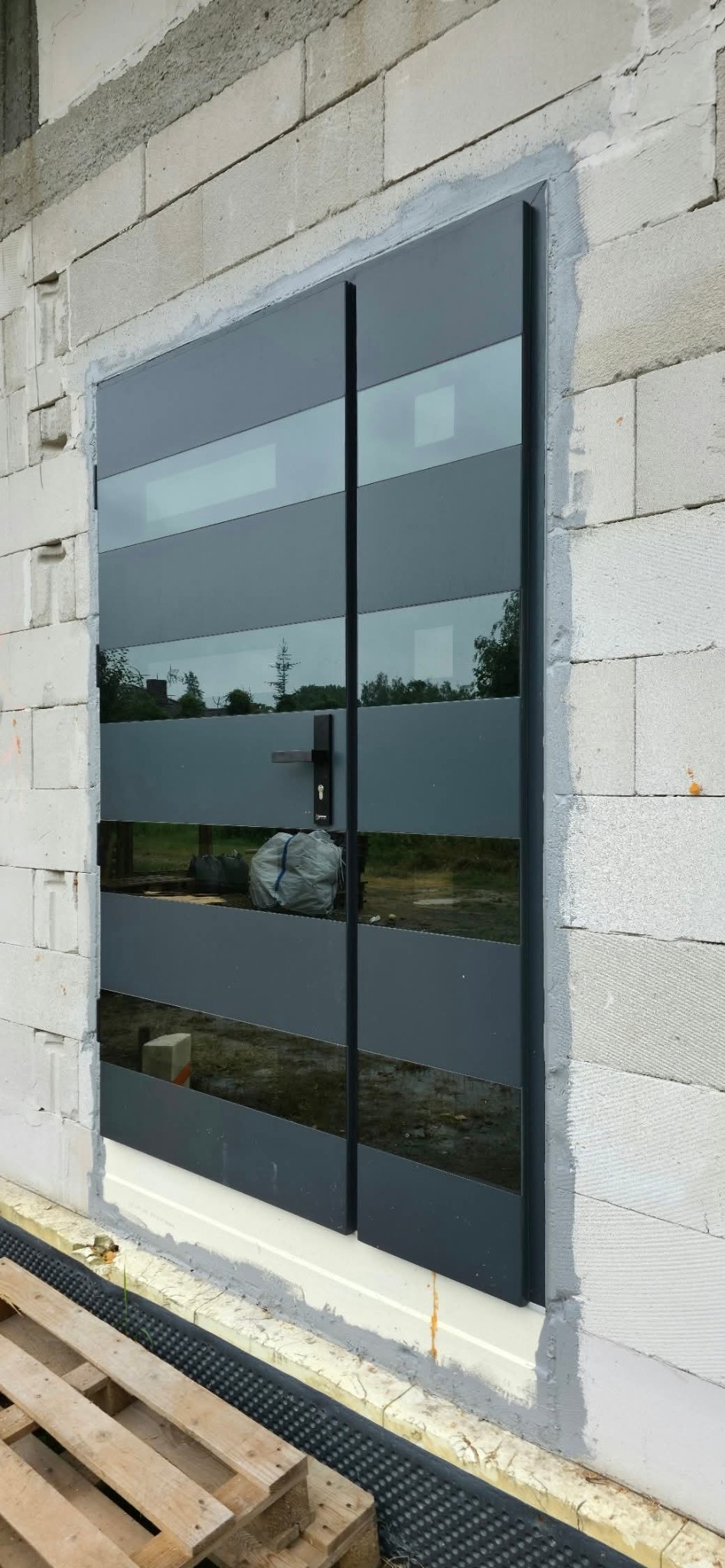 Nowoczesne, dwuskrzydłowe drzwi zewnętrzne z poziomymi przeszkleniami, zamontowane w surowym murze z betonu komórkowego. Widoczny fragment palety drewnianej.