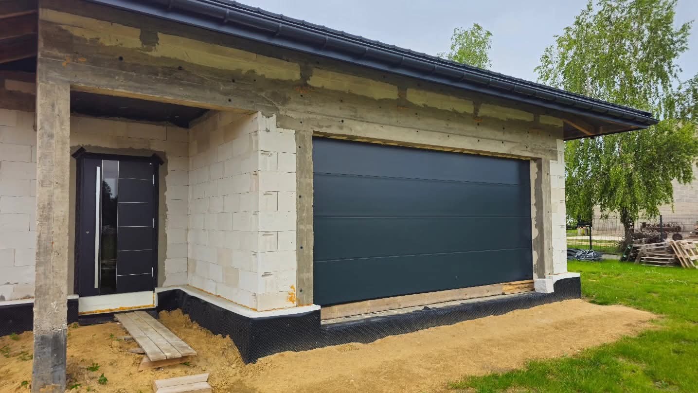 Nowoczesna, grafitowa brama garażowa segmentowa i drzwi wejściowe w surowym stanie budynku z białego bloczka, widok z zewnątrz, dzień.