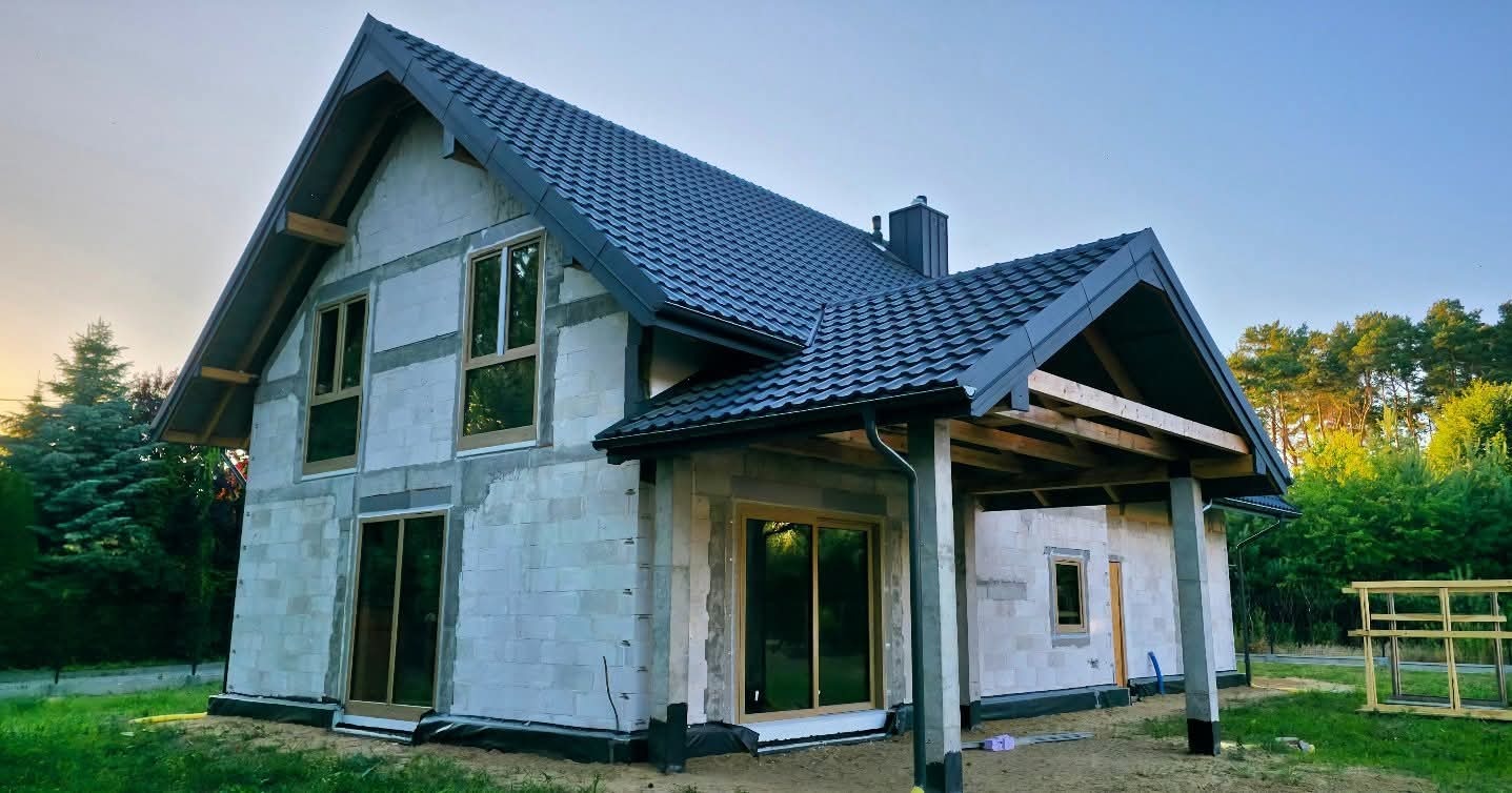 Surowy dom jednorodzinny z szarą dachówką, widoczne okna z drewnianą ramą i ściany z białego bloczka. Budynek w trakcie budowy, z elementami konstrukcji zadaszenia.