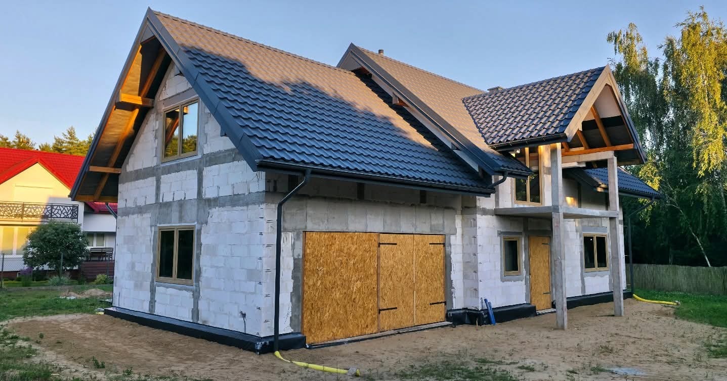 Dom w trakcie budowy z widoczną konstrukcją dachu, oknami i tymczasowo zamkniętą bramą garażową. Ściany z białych bloczków, czarna dachówka. Widok z zewnątrz, w słoneczny dzień.