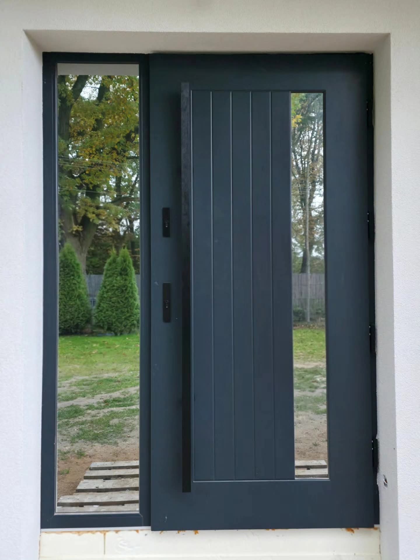 Nowoczesne, grafitowe drzwi zewnętrzne z bocznym przeszkleniem, widok na ogród z tujami. Minimalistyczny design, pionowe panele, naturalne światło.