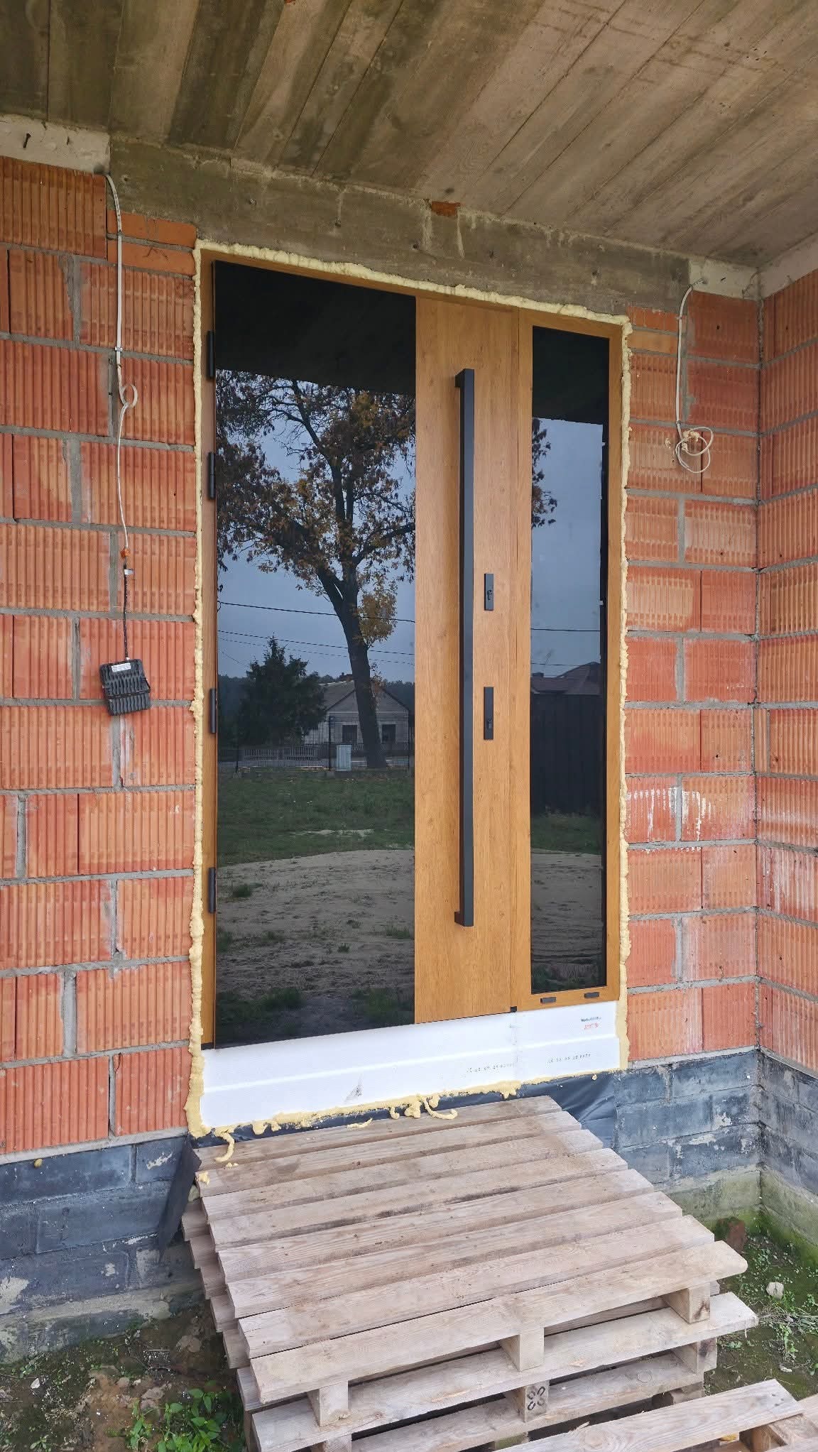 Nowoczesne drzwi zewnętrzne z długim, czarnym uchwytem, zamontowane w surowym murze z czerwonej cegły. Widoczne pianka montażowa i paleta drewniana.