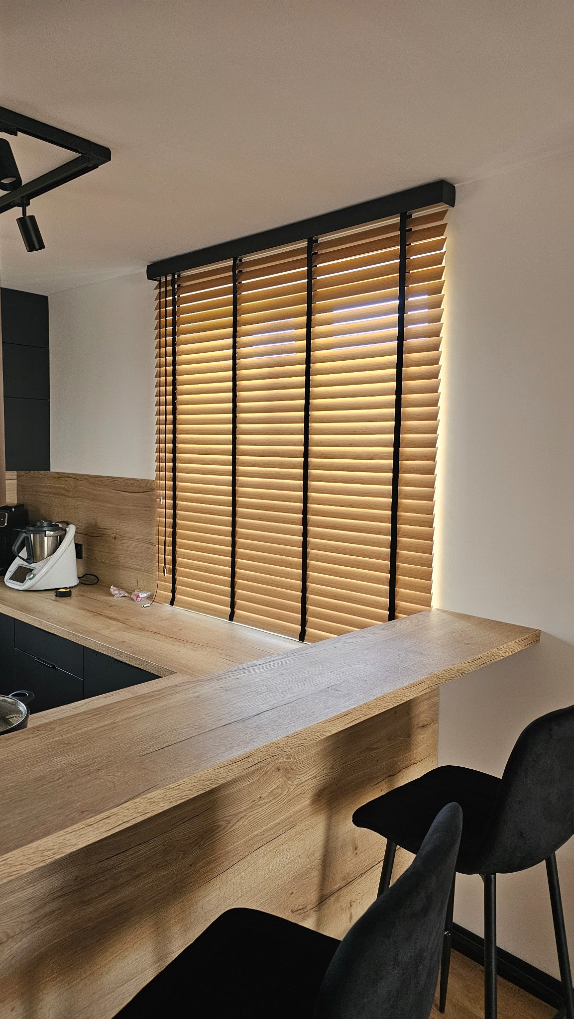 Drewniane żaluzje poziome w kuchni z blatem i krzesłami barowymi. Naturalne światło podkreśla strukturę drewna. Minimalistyczny design.