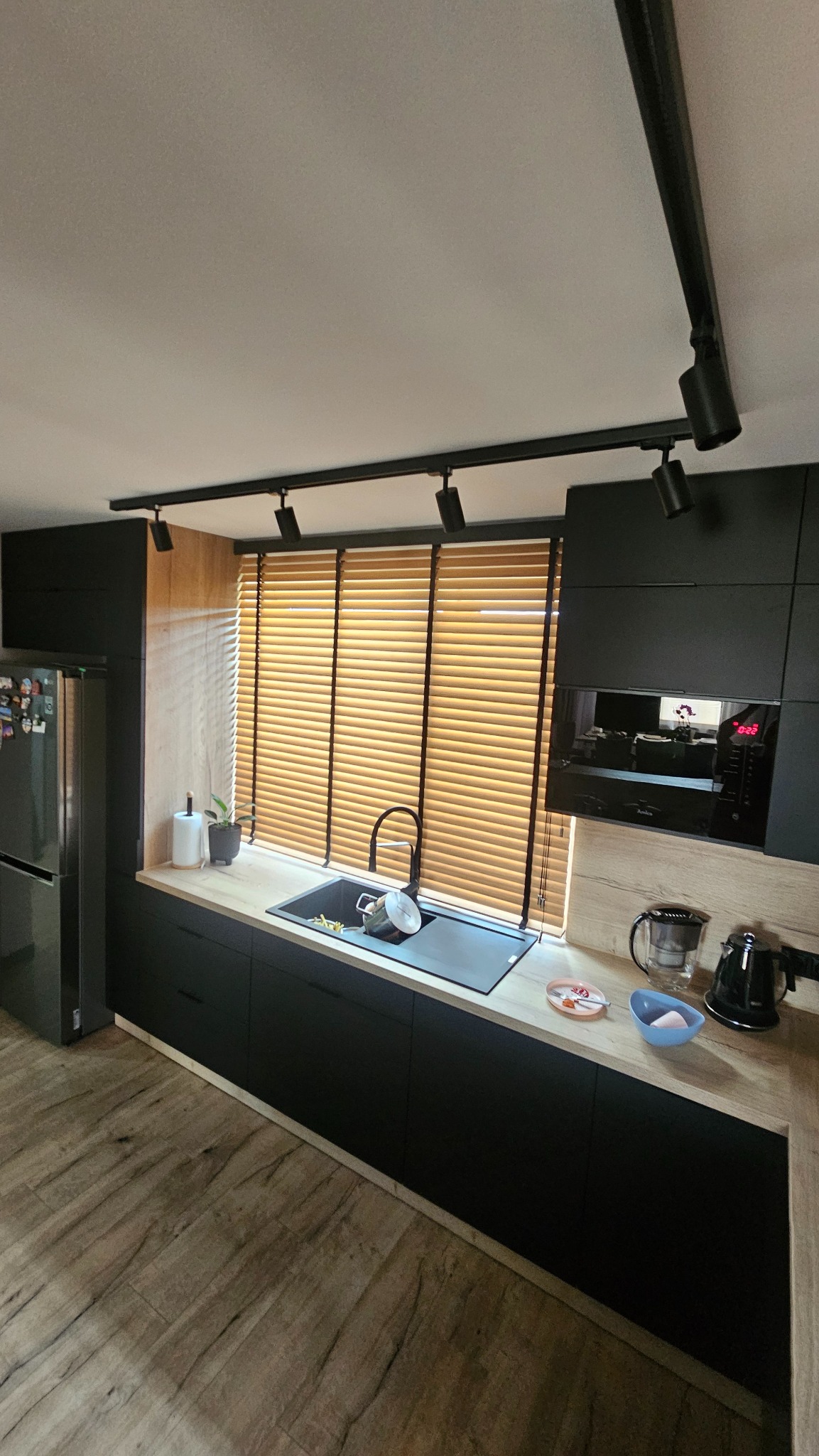Nowoczesna kuchnia z drewnianymi żaluzjami poziomymi w oknie nad zlewem. Czarna armatura i oświetlenie szynowe podkreślają minimalistyczny design.