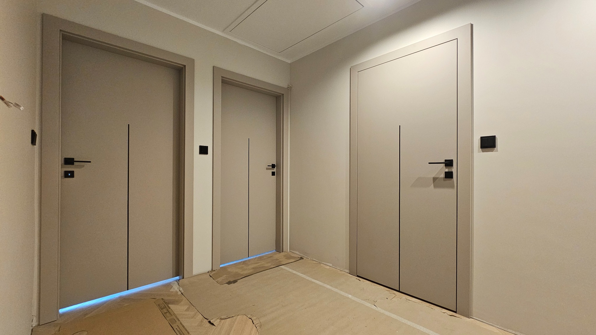 Trzy beżowe drzwi wewnętrzne z czarnymi klamkami i pionowymi frezami, zamontowane w jasnym korytarzu. Podłoga zabezpieczona kartonem.