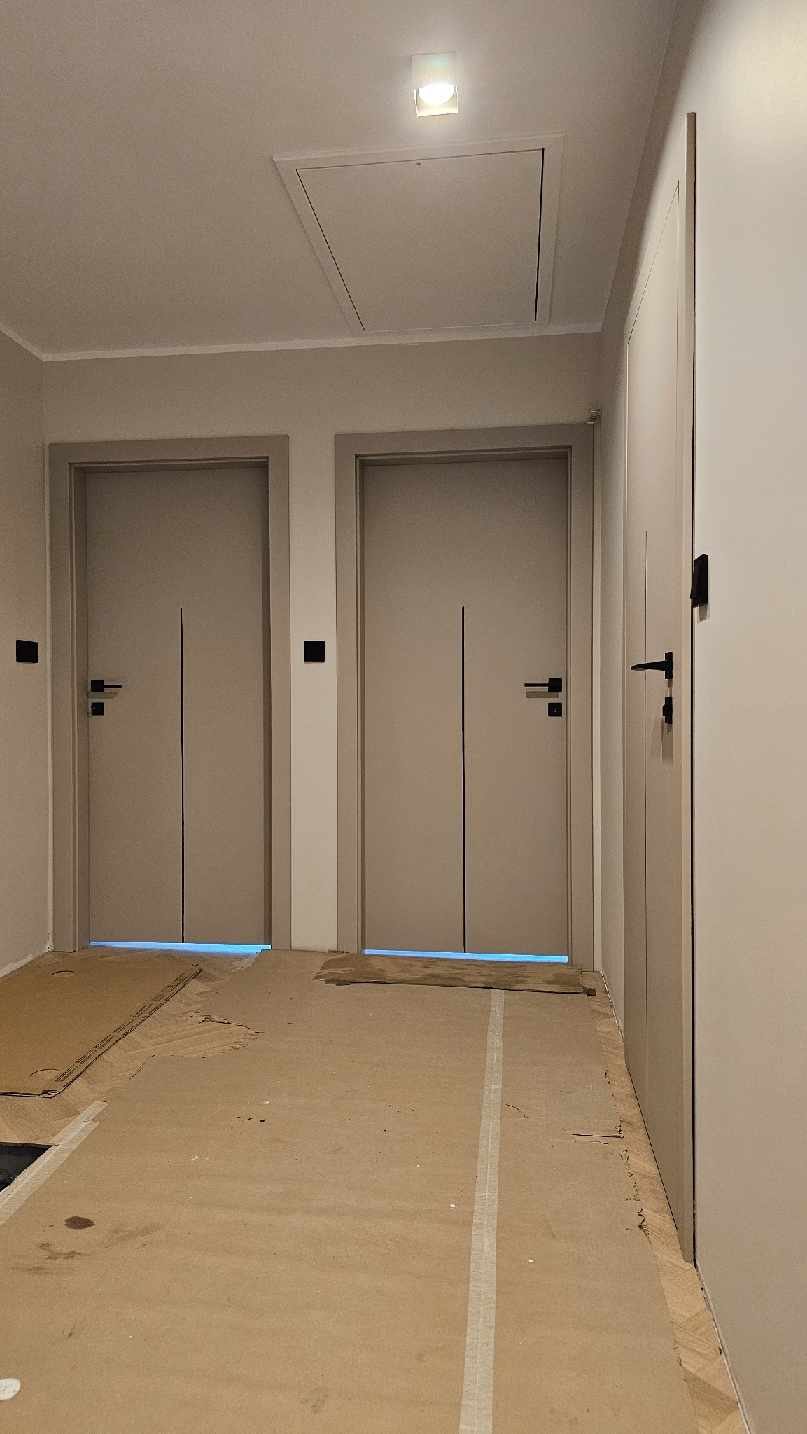 Jasne, minimalistyczne drzwi wewnętrzne z czarnymi klamkami, widoczne w korytarzu podczas prac wykończeniowych, zabezpieczona podłoga.