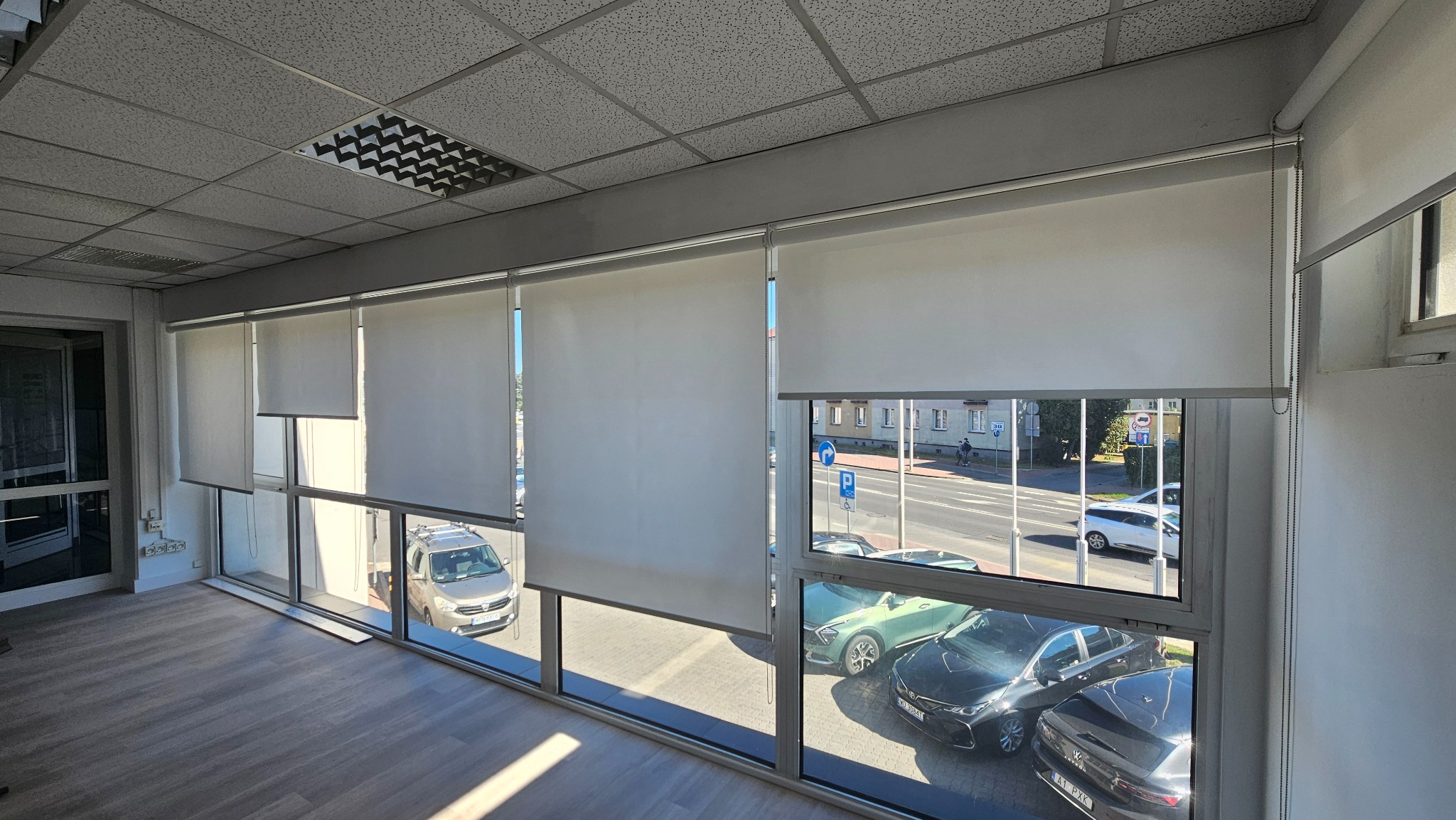 Białe rolety zwijane zamontowane w oknach biura, widok na ulicę z samochodami. Rolety częściowo opuszczone, wpadające światło dzienne.