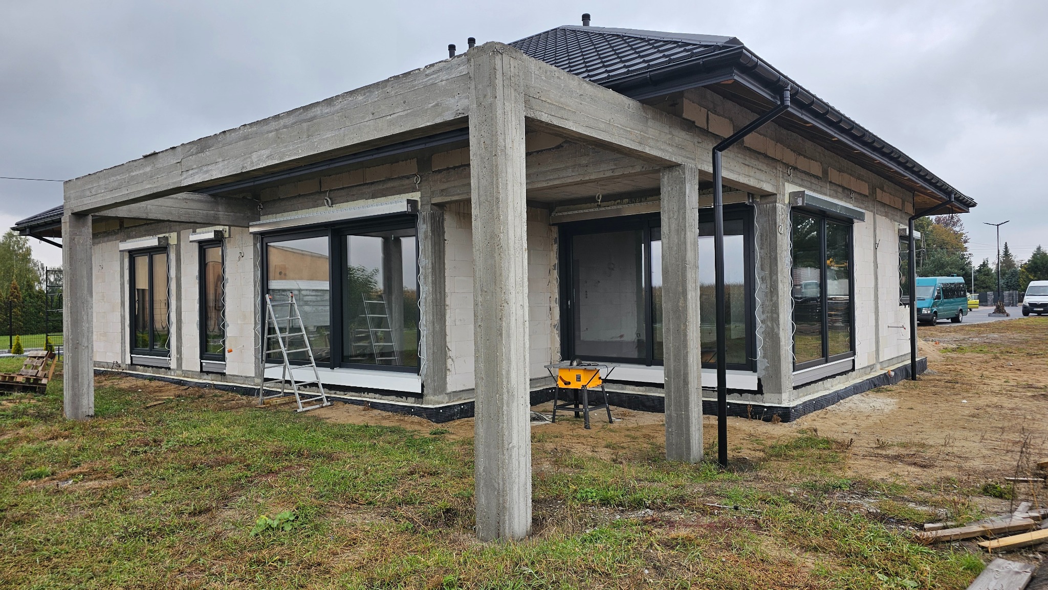 Surowy stan budynku z zamontowanymi oknami i roletami zewnętrznymi. Widoczny szary beton, drabina i narzędzia. Budowa w toku, pochmurne niebo w tle.