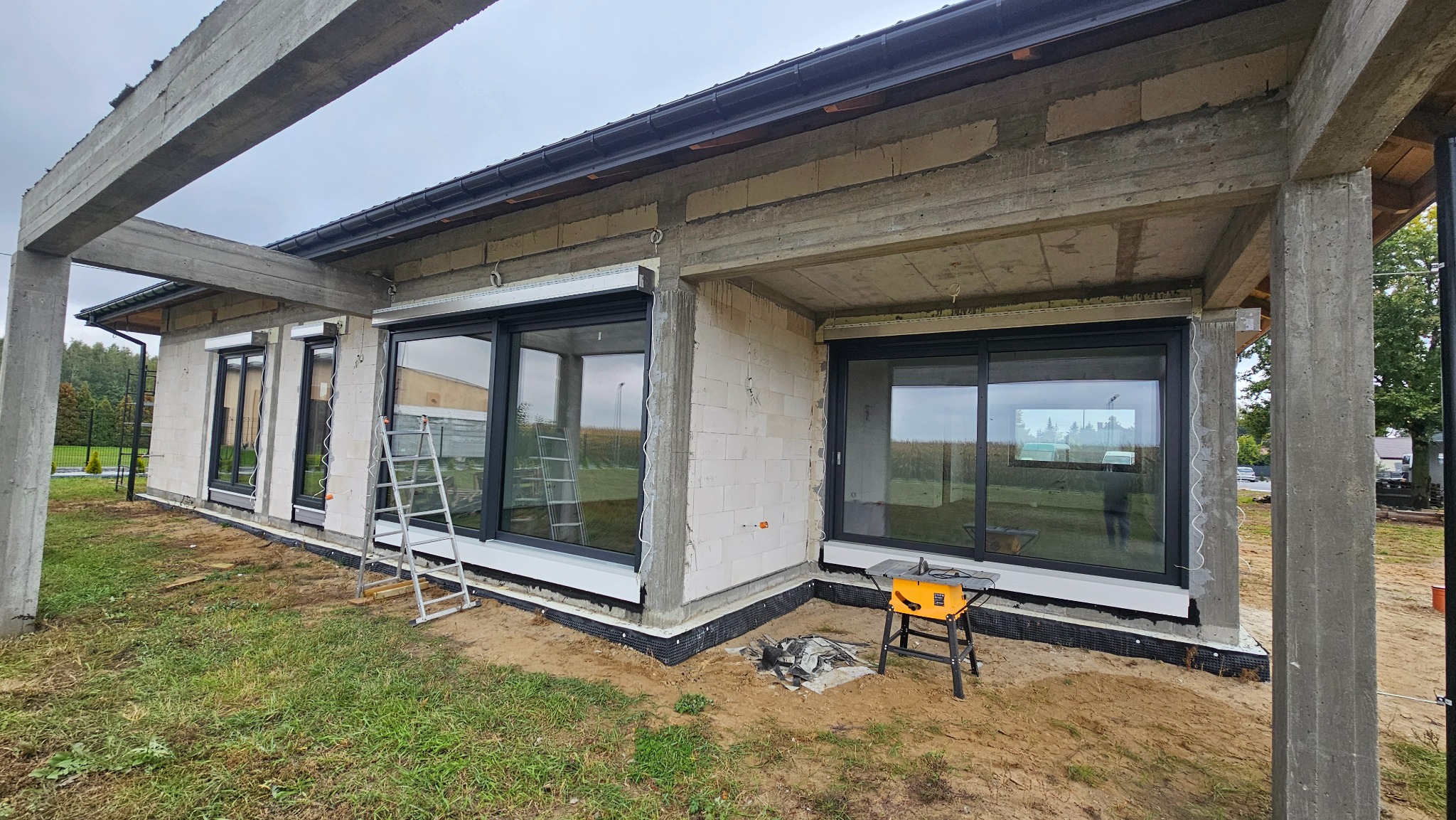 Surowy stan budynku z zamontowanymi oknami z czarnymi ramami i roletami zewnętrznymi. Na zewnątrz drabina i stół roboczy z narzędziami.