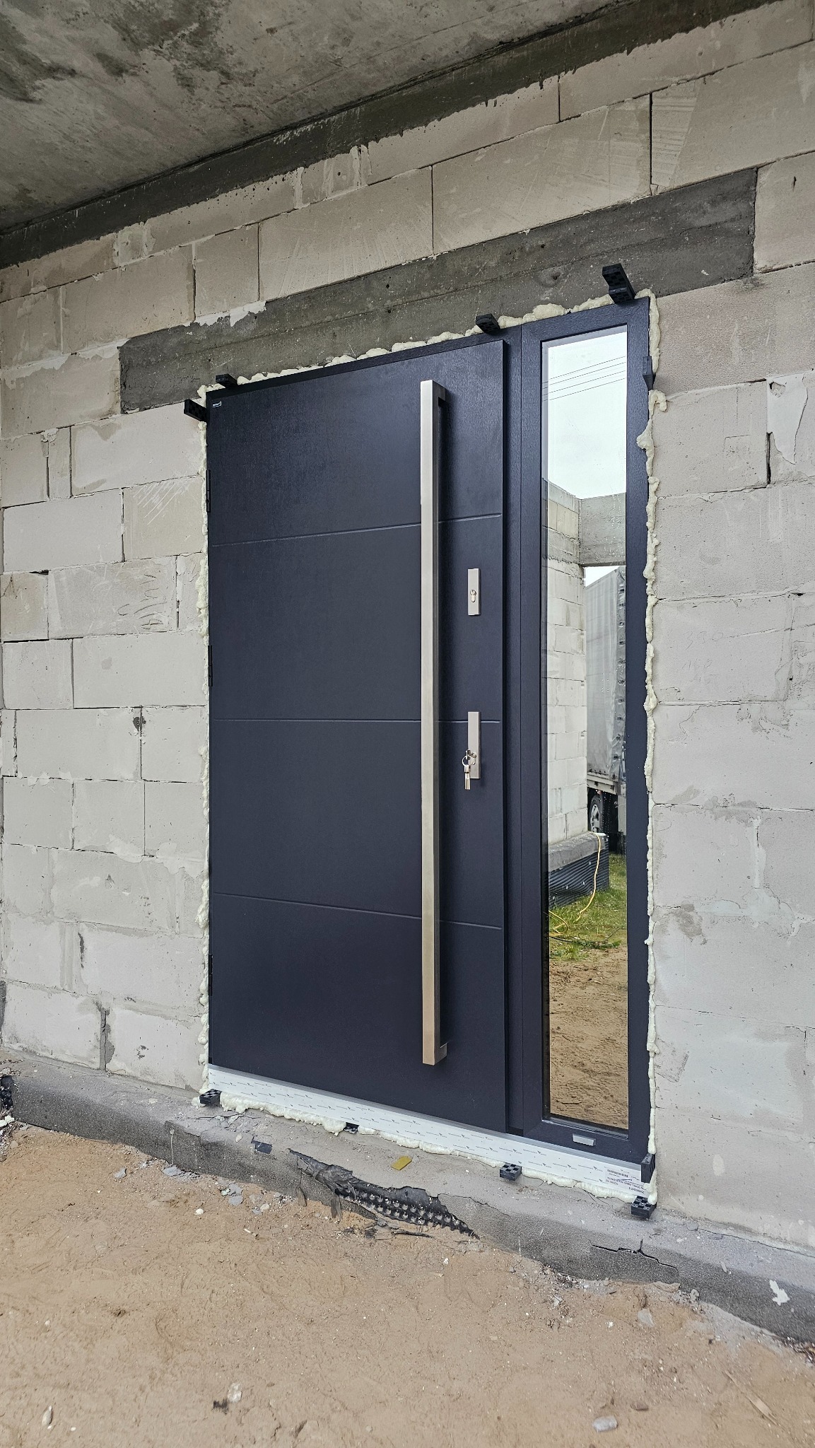Ciemne drzwi zewnętrzne z długim uchwytem, zamontowane w surowym murze z betonu komórkowego. Widoczna pianka montażowa. Usługa montażu w Płocku.
