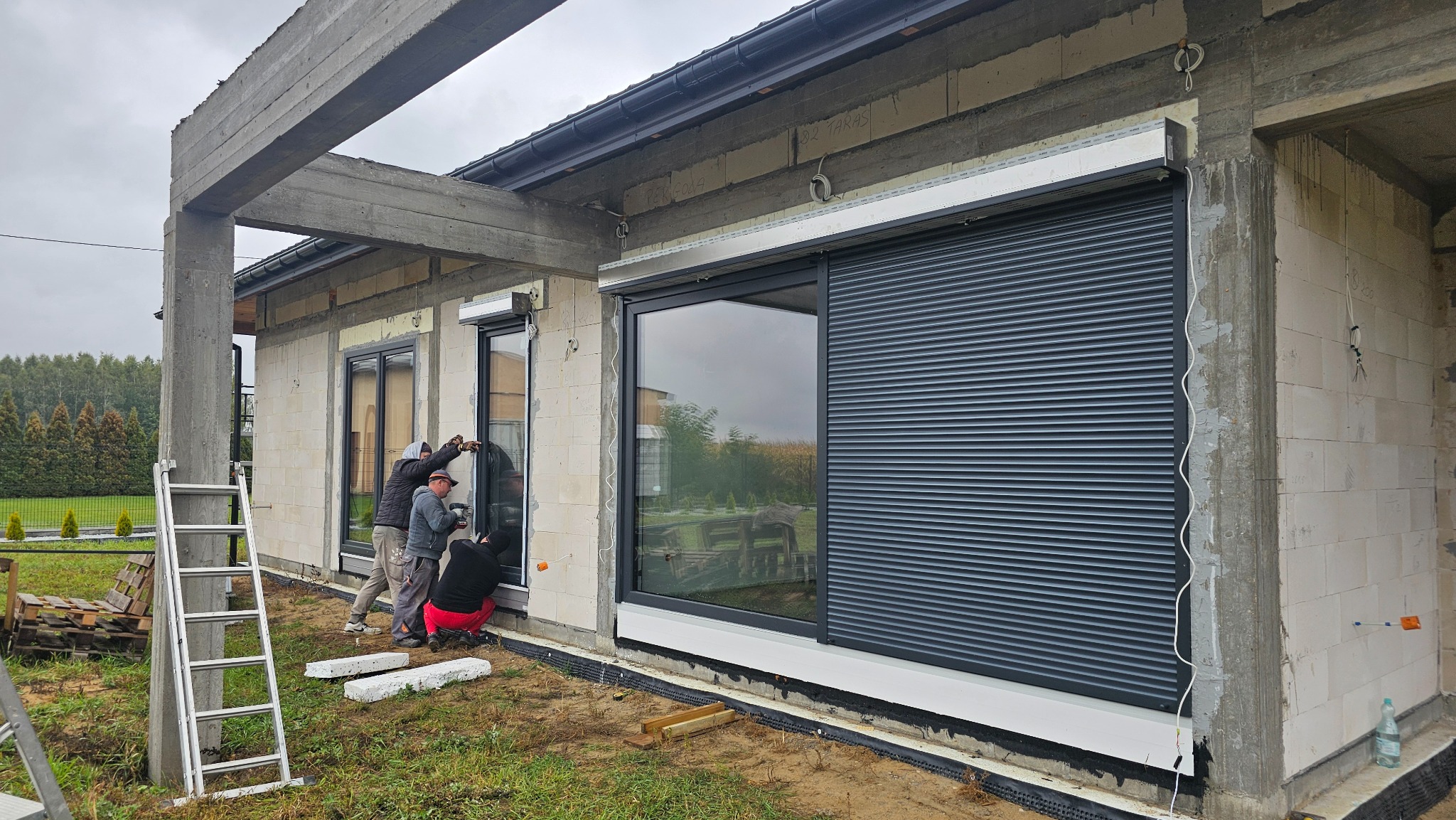 Montaż okien i rolet zewnętrznych w surowym budynku. Widoczne okna z zamontowanymi roletami, drabina i trzech monterów podczas pracy. Budynek w trakcie budowy.