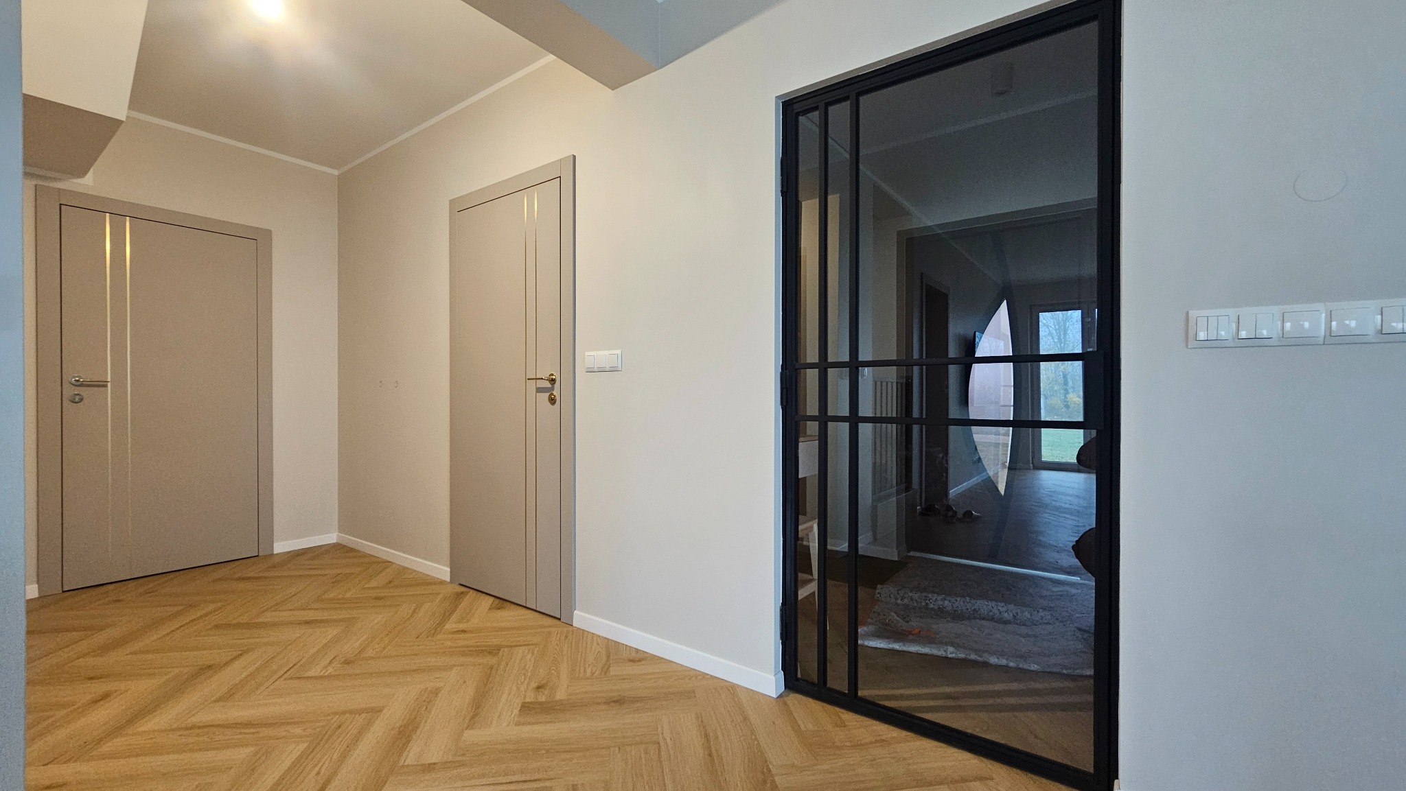 Nowoczesne drzwi wewnętrzne z czarną ramą i szkłem, widoczne na tle parkietu w jodełkę i dwóch beżowych drzwi. Minimalistyczny design, jasne wnętrze.