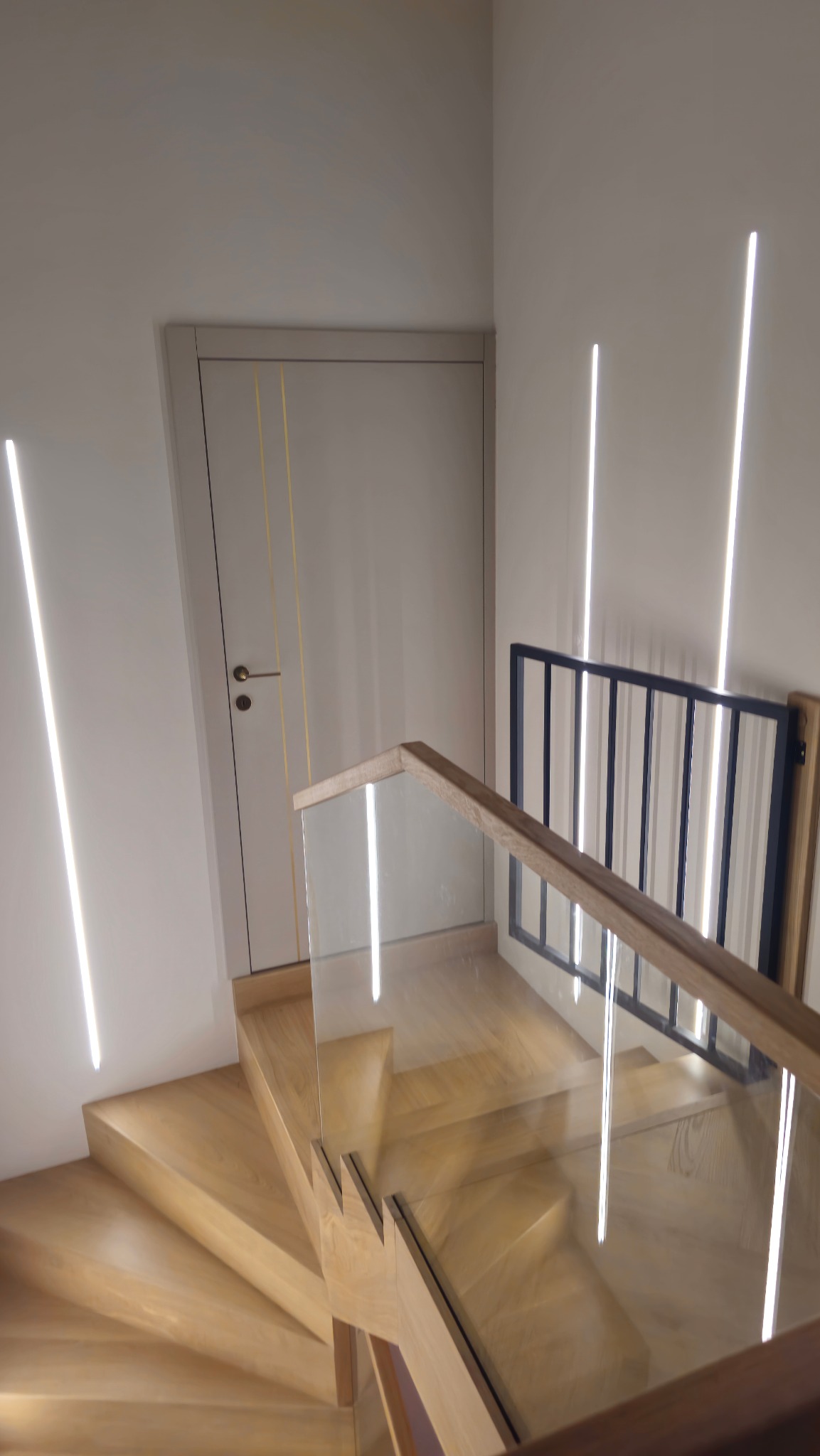 Nowoczesne schody drewniane z balustradą ze szkła i drewna, oświetlone pionowymi lampami LED na ścianie. Widoczny fragment drzwi wewnętrznych z delikatnymi zdobieniami.