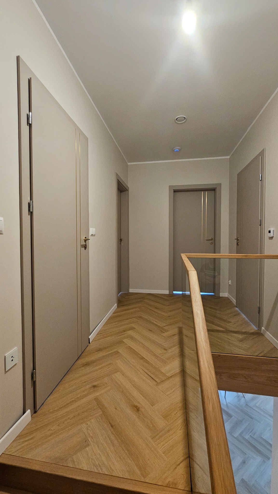 Korytarz z parkietem w jodełkę i beżowymi drzwiami. Widoczne trzy pary drzwi, oświetlenie sufitowe i drewniana balustrada. Minimalistyczny design.