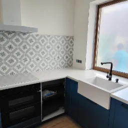 Niebieska kuchnia z białym blatem, wzorzystymi płytkami nad blatem i białym zlewem farmerskim pod oknem z folią ochronną.