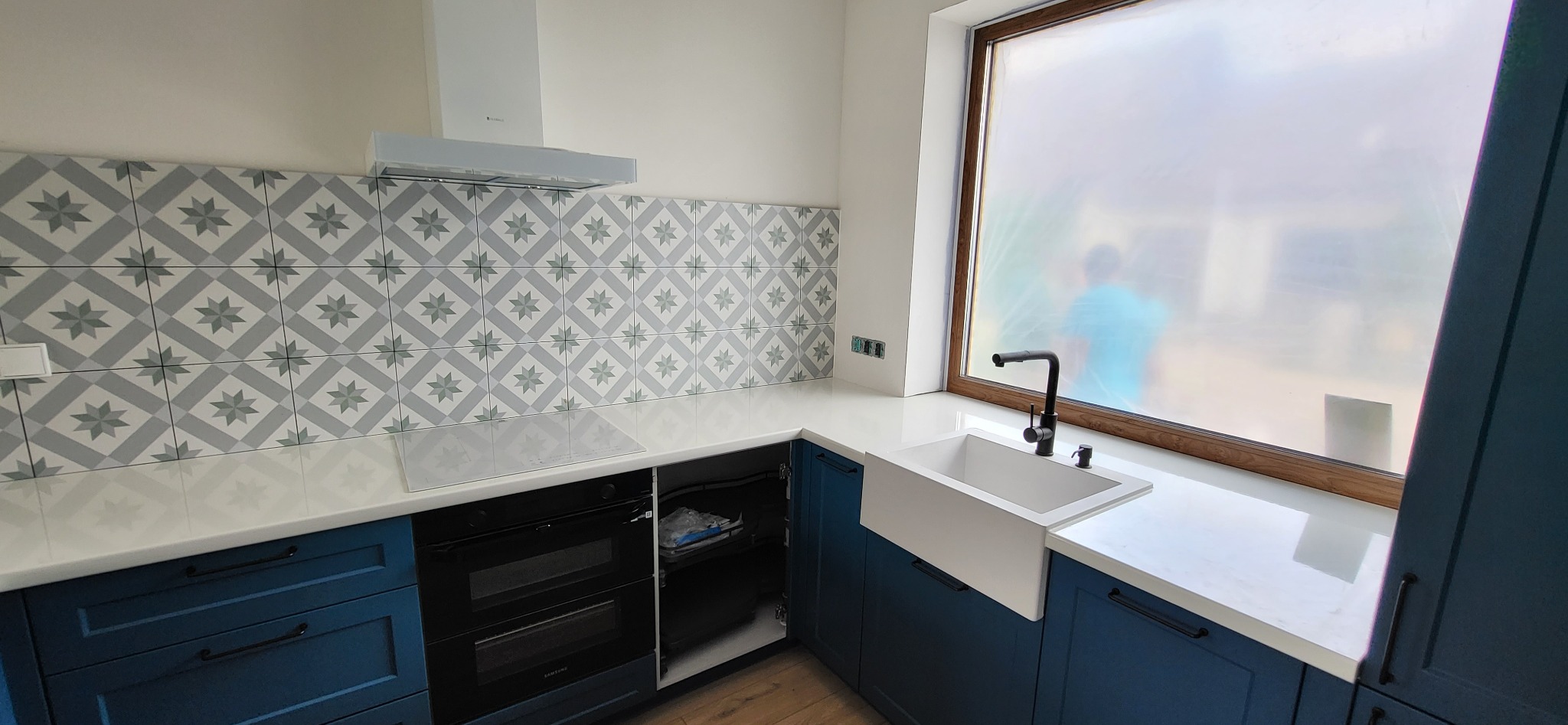 Niebieska kuchnia z białym blatem, wzorzystymi płytkami nad blatem i białym zlewem farmerskim pod oknem z folią ochronną.