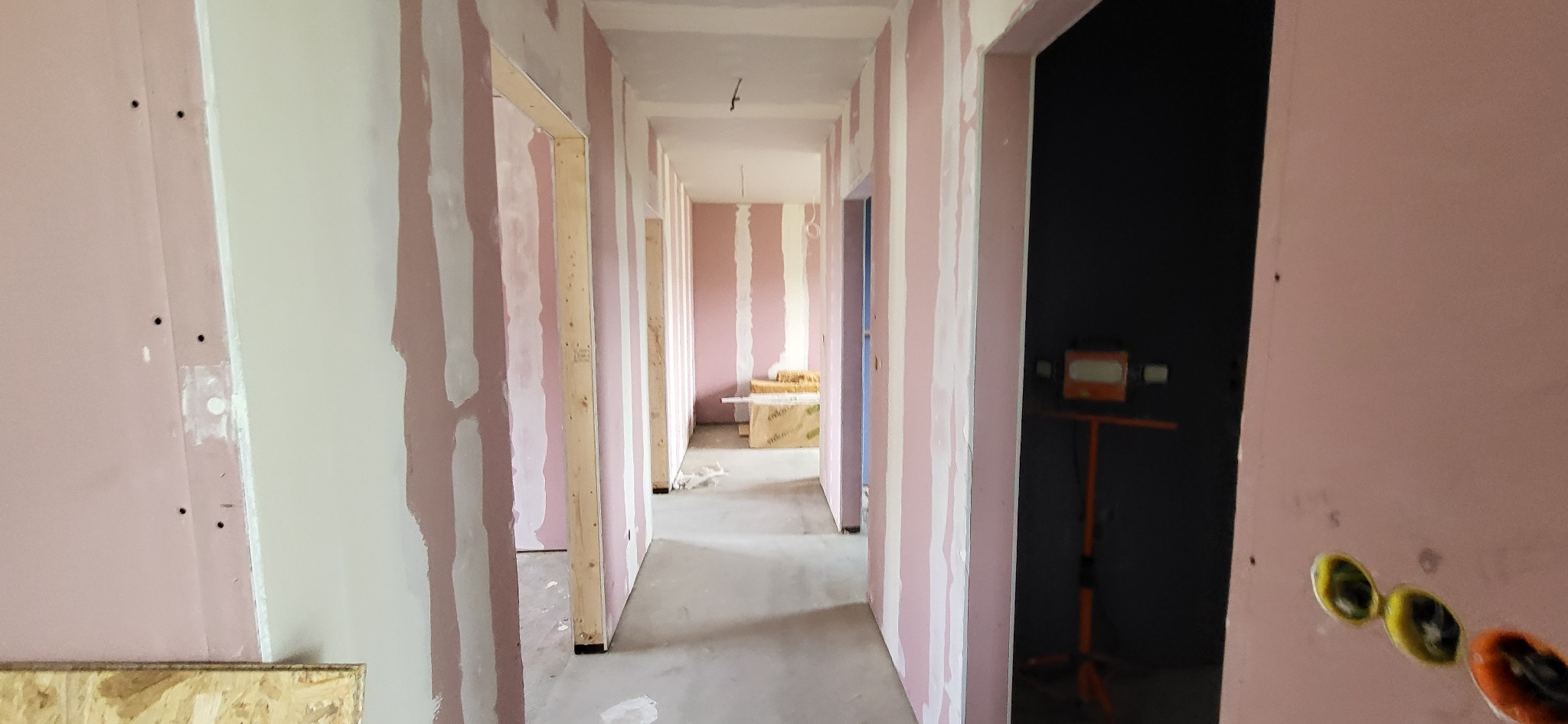 Korytarz w trakcie remontu, widoczne ściany z płyt gipsowo-kartonowych przed szpachlowaniem, z widocznymi łączeniami i otworami po wkrętach, surowe drewniane futryny drzwiowe, w tle materiały...