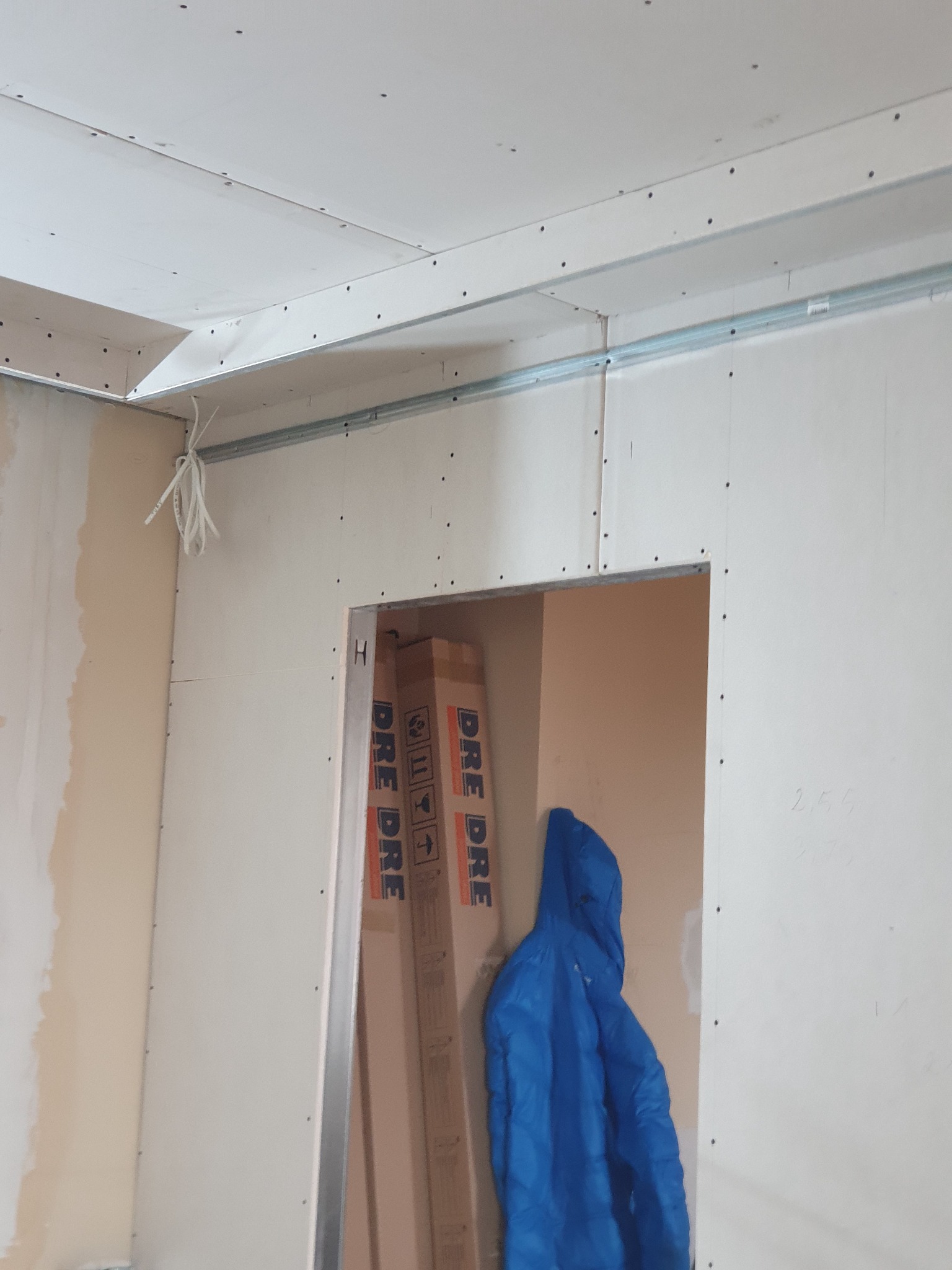 Wnętrze w trakcie montażu płyt gipsowo-kartonowych na ścianach i suficie, widoczne profile metalowe, przewody elektryczne oraz kartony drzwi w otworze drzwiowym.