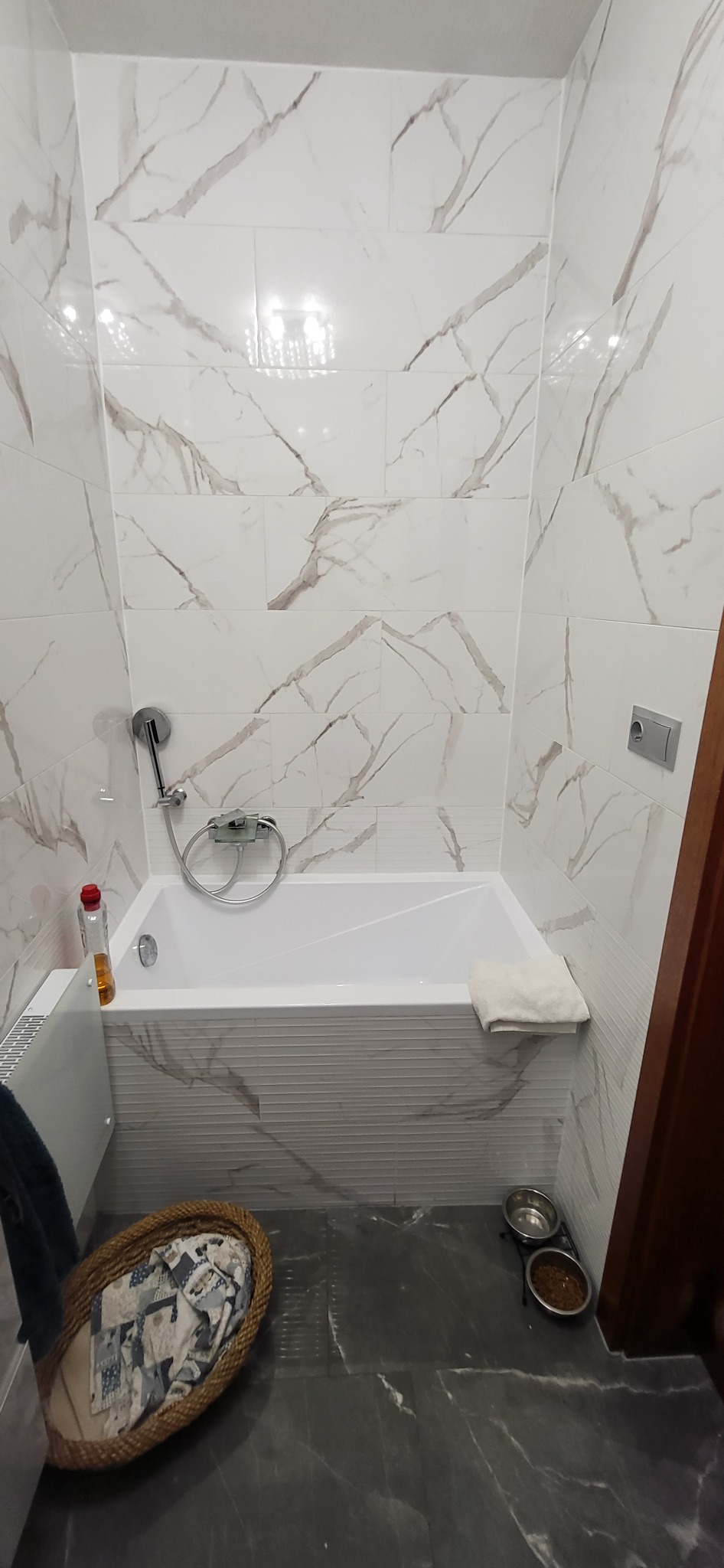 Wykończona łazienka z białą wanną obudowaną płytkami, ściany wyłożone białymi płytkami imitującymi marmur, szara marmurowa podłoga, widoczne miski dla zwierząt i kosz na pranie.