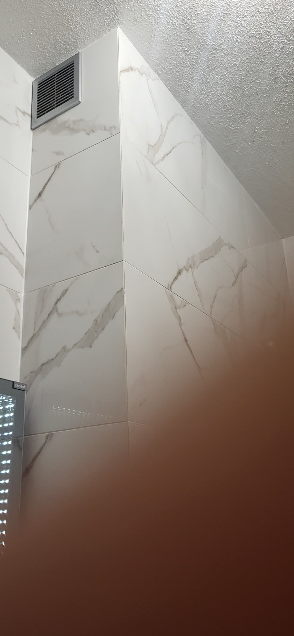 Wykończona ściana w łazience z białych, marmurkowych płytek ceramicznych, widoczna kratka wentylacyjna w górnym rogu.