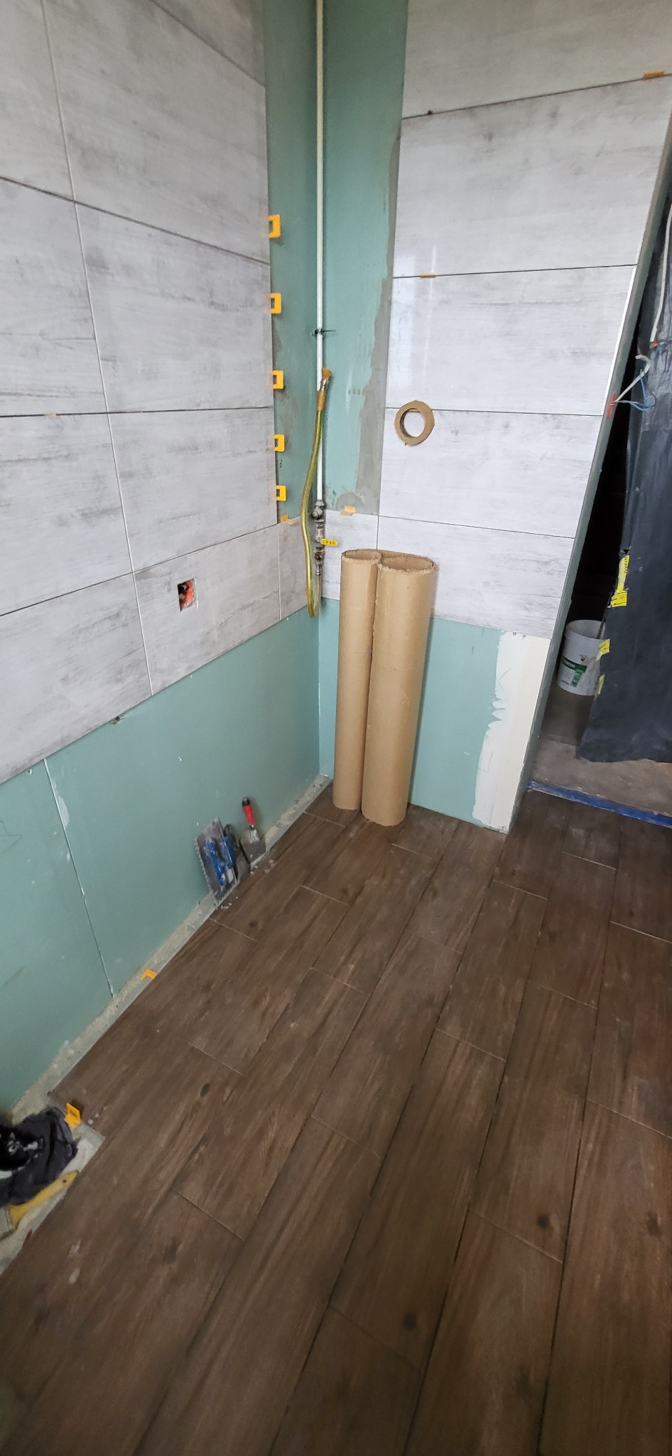 Wnętrze łazienki w trakcie remontu: częściowo ułożone płytki imitujące drewno na podłodze, szare płytki ścienne z żółtymi krzyżykami dystansowymi, widoczne rury, narzędzia i kartonowe tuby.