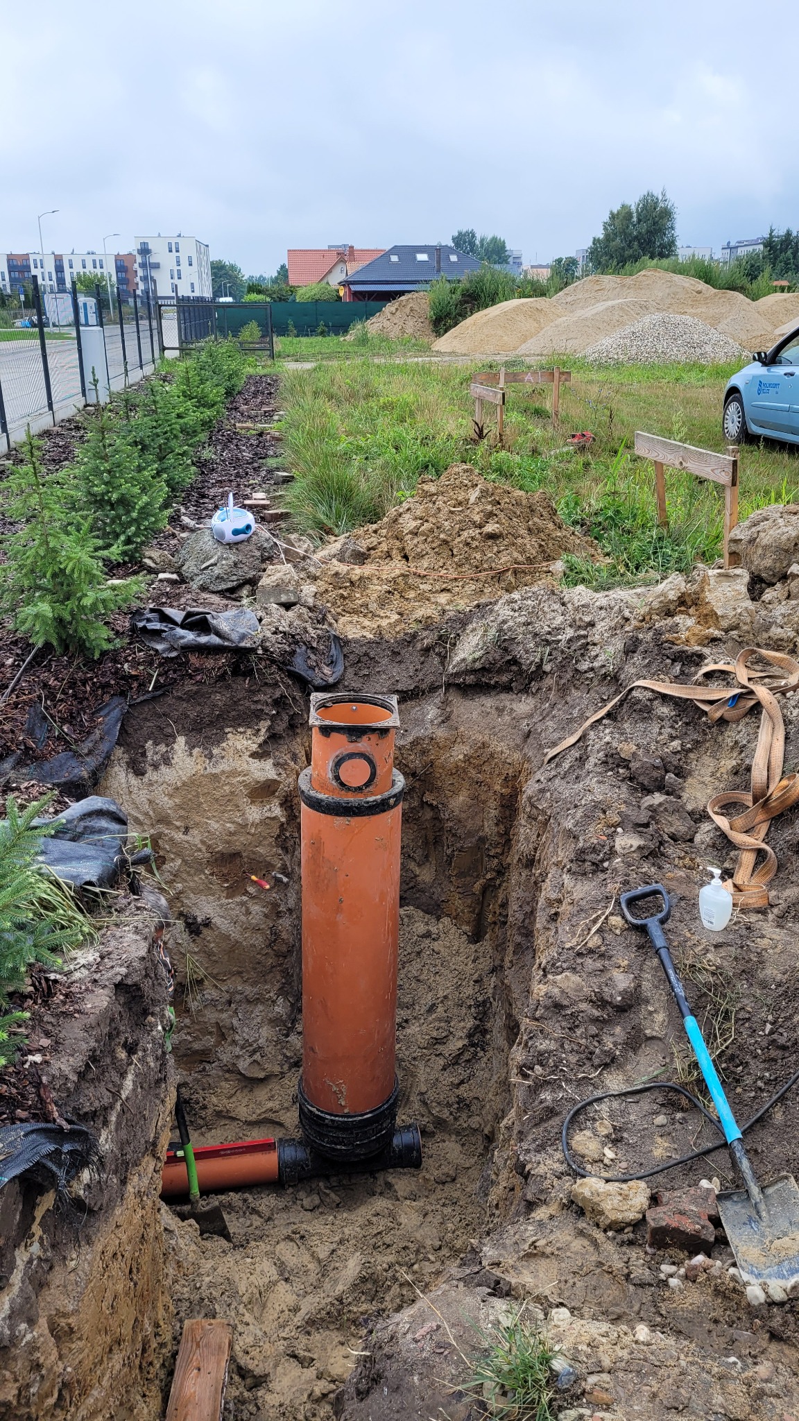 Pomarańczowa rura kanalizacyjna pionowo umieszczona w wykopie ziemnym, z widocznymi przyłączami, na tle trawy, drzew i budynków.