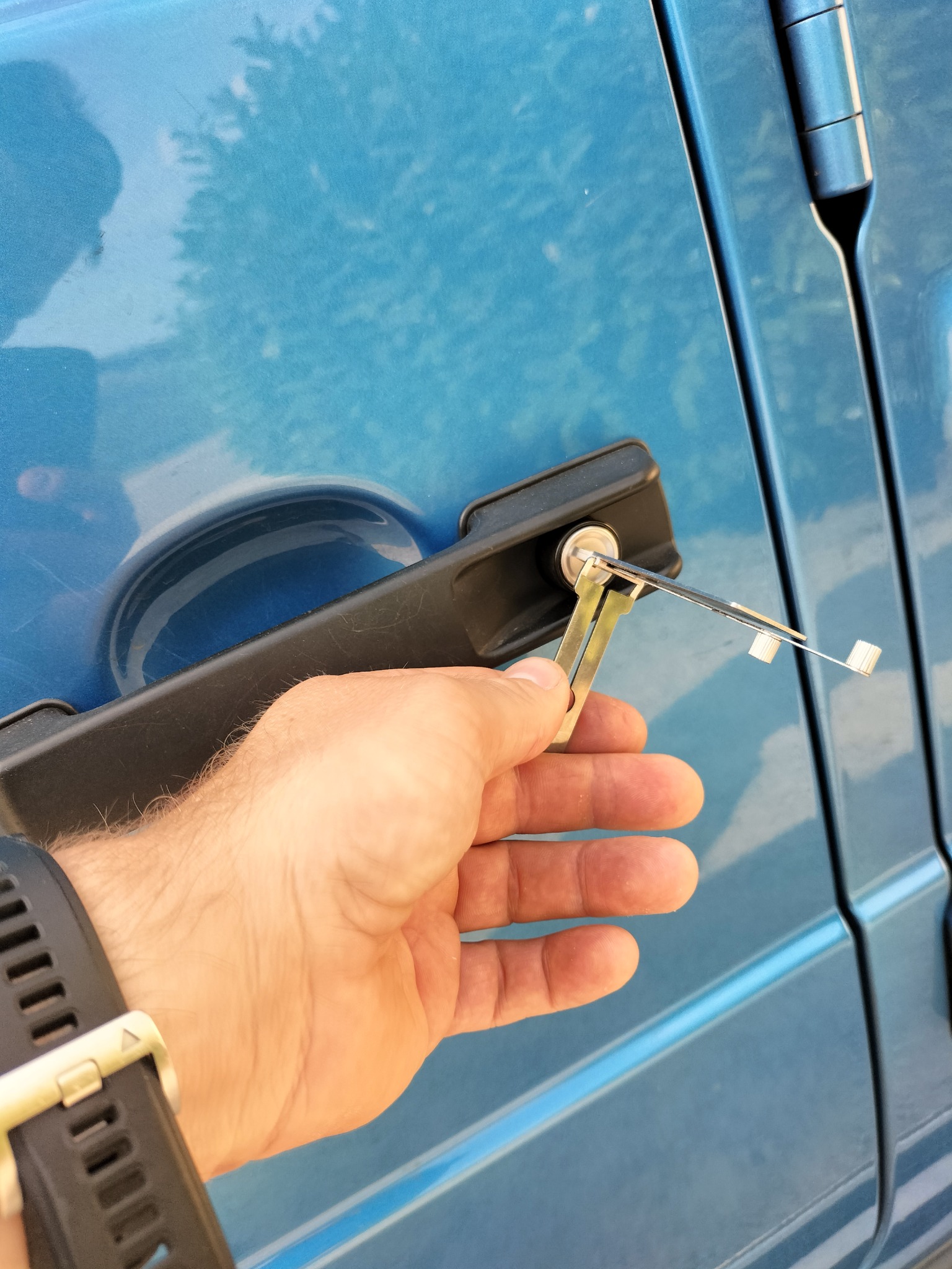 Otwieranie zamka w drzwiach samochodu za pomocą wytrychów, widoczna ręka z zegarkiem na tle niebieskiego lakieru.