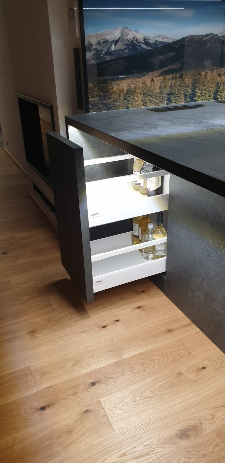 Nowoczesna kuchnia z wysuwaną szafką typu cargo marki Blum, podświetloną od wewnątrz, wypełnioną butelkami i słoikami, z blatem imitującym kamień i drewnianą podłogą, widok na fototapetę z górskim...