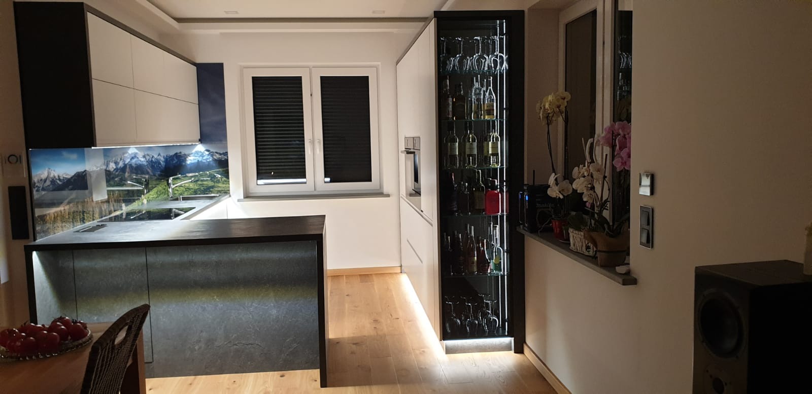 Nowoczesna kuchnia z wyspą, białymi szafkami i szklanym panelem z motywem górskim, podświetlana szafka z kolekcją alkoholi i kieliszków, drewniana podłoga.