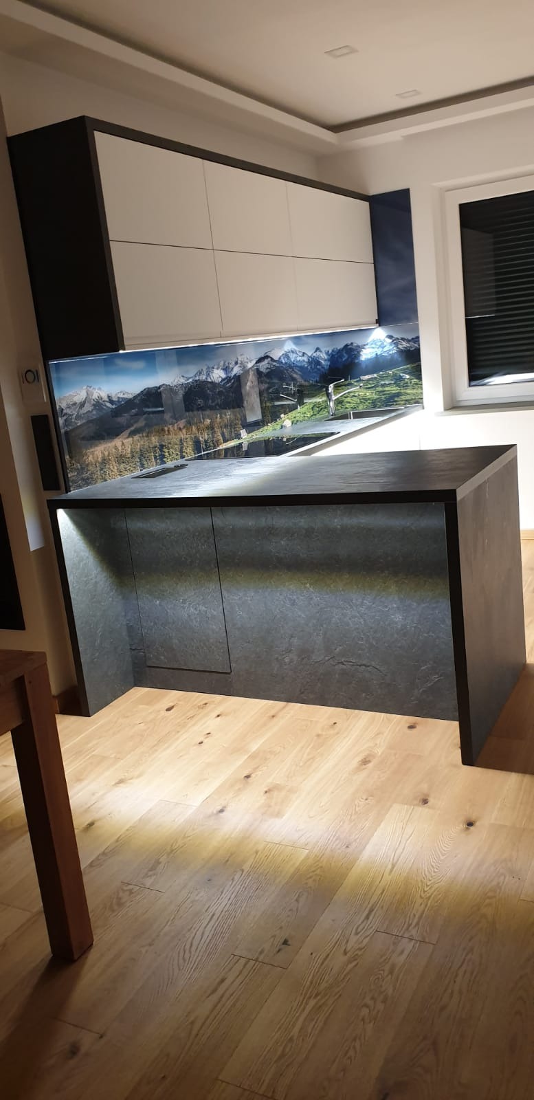 Wyspa kuchenna z szarym blatem i frontami imitującymi kamień, podświetlona od dołu, z górnymi szafkami w kolorze białym i czarnym oraz panelem szklanym z motywem górskim.