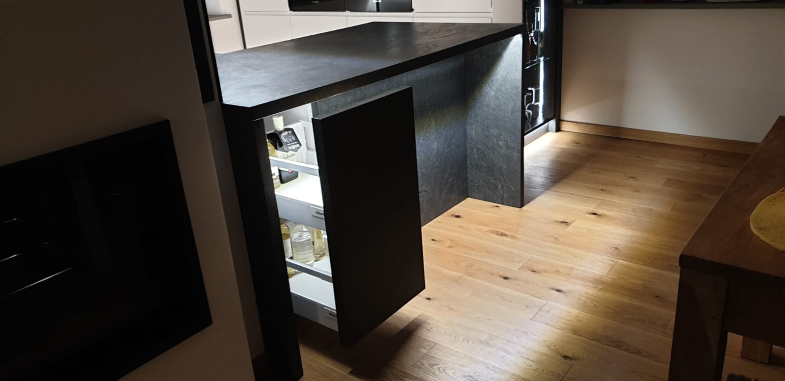 Nowoczesna wyspa kuchenna z ciemnym blatem i frontami, z otwartą szufladą chłodziarki wypełnioną butelkami i słoikami, oświetlona od wewnątrz, na podłodze drewniany parkiet.