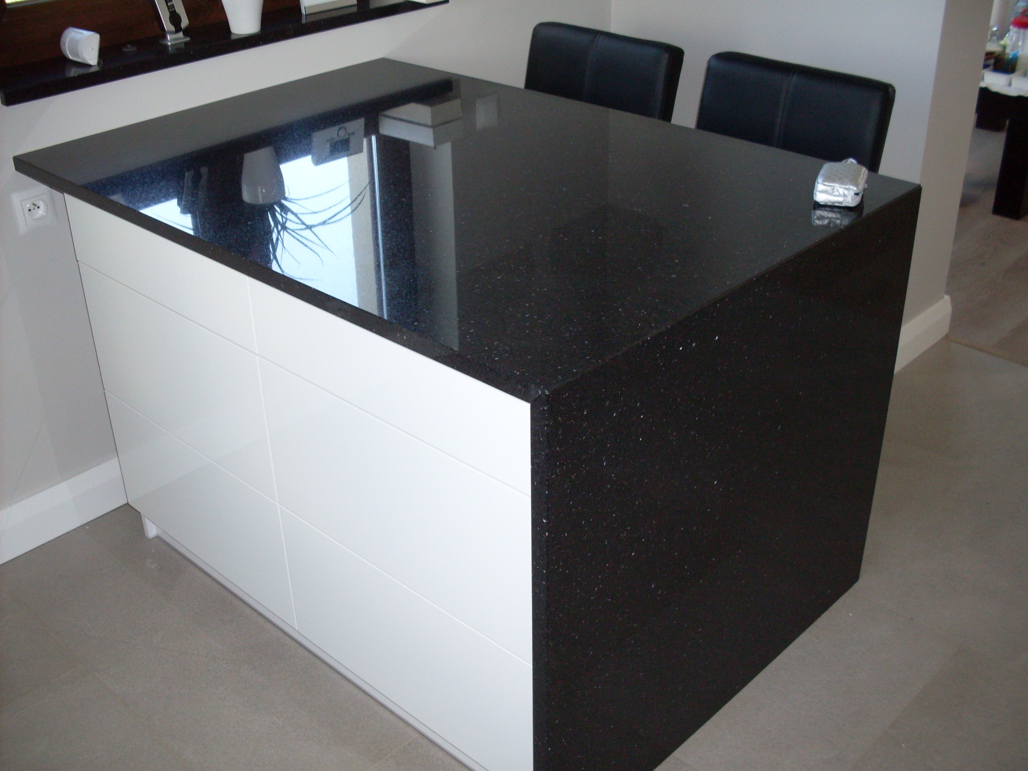 Wyspa kuchenna z czarnym, błyszczącym blatem z drobinkami i białymi, poziomymi frontami szafek, uzupełniona o dwa czarne krzesła barowe w tle.