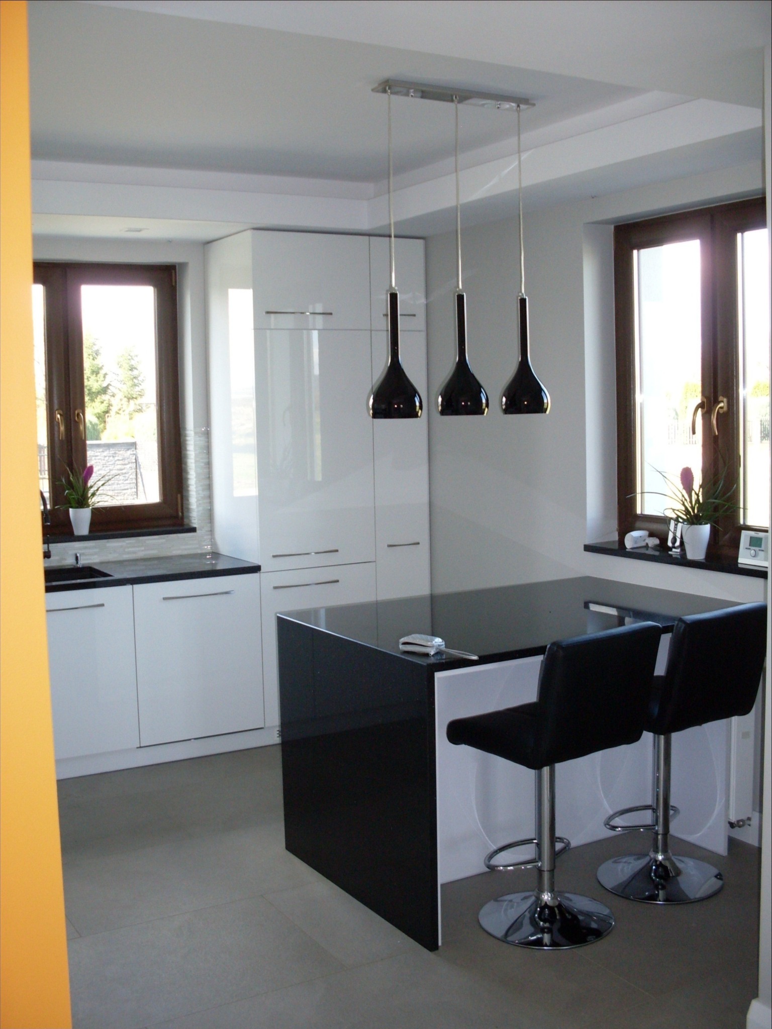 Nowoczesna kuchnia w bieli i czerni z wyspą, trzema wiszącymi lampami w kształcie łezki, dwoma chromowanymi hokerami i roślinami w doniczkach przy oknach.