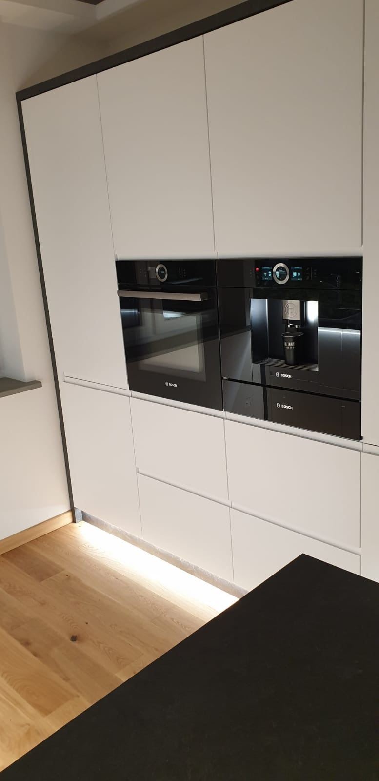 Nowoczesna zabudowa kuchenna z białymi frontami, wbudowanym piekarnikiem i ekspresem do kawy marki Bosch, podświetleniem LED przy podłodze i fragmentem czarnego blatu wyspy.
