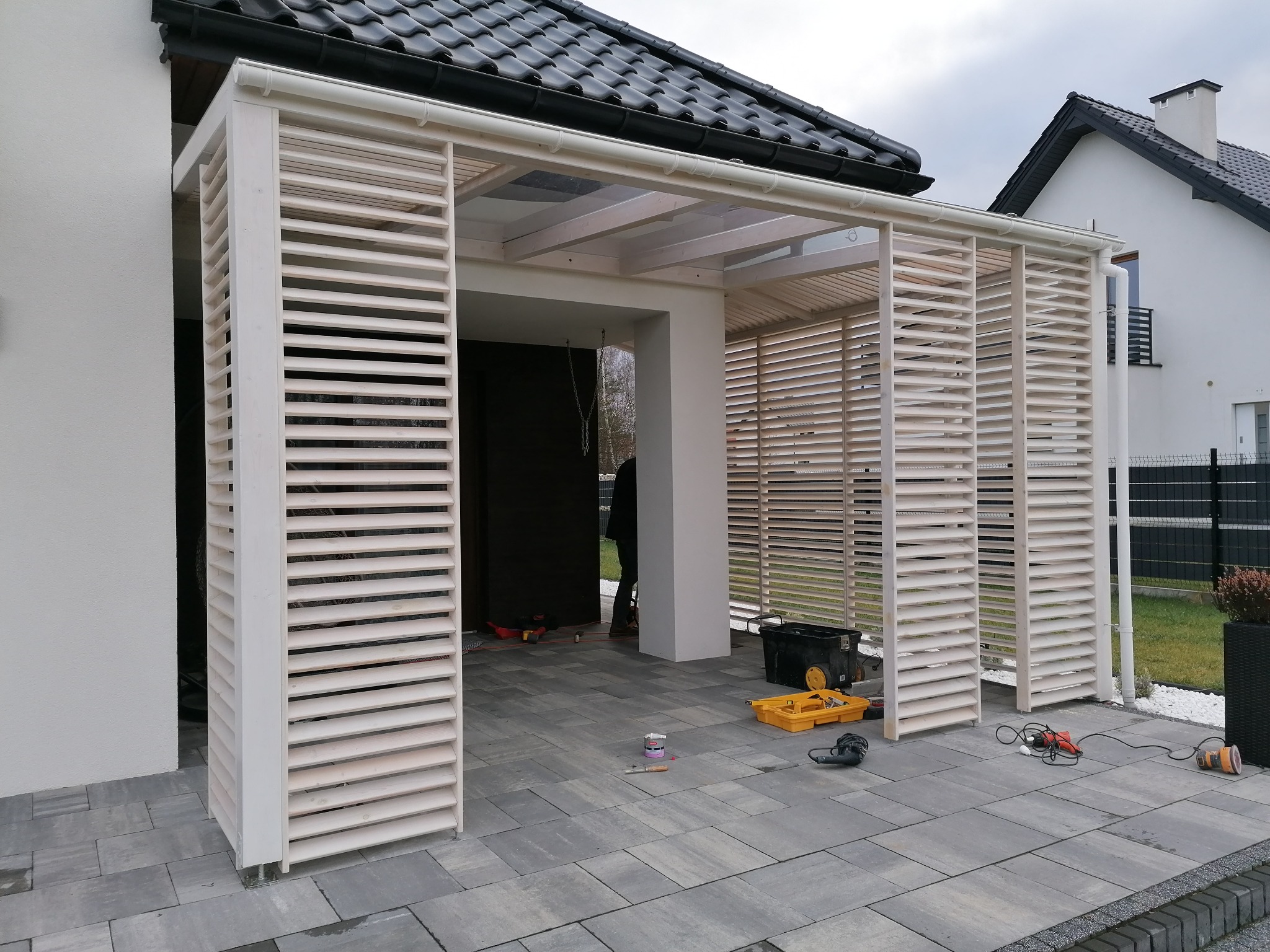 Drewniana pergola z lamelami w kolorze jasnego beżu, częściowo zadaszona przeźroczystym tworzywem, na szarej kostce brukowej, widoczne narzędzia i akcesoria montażowe.