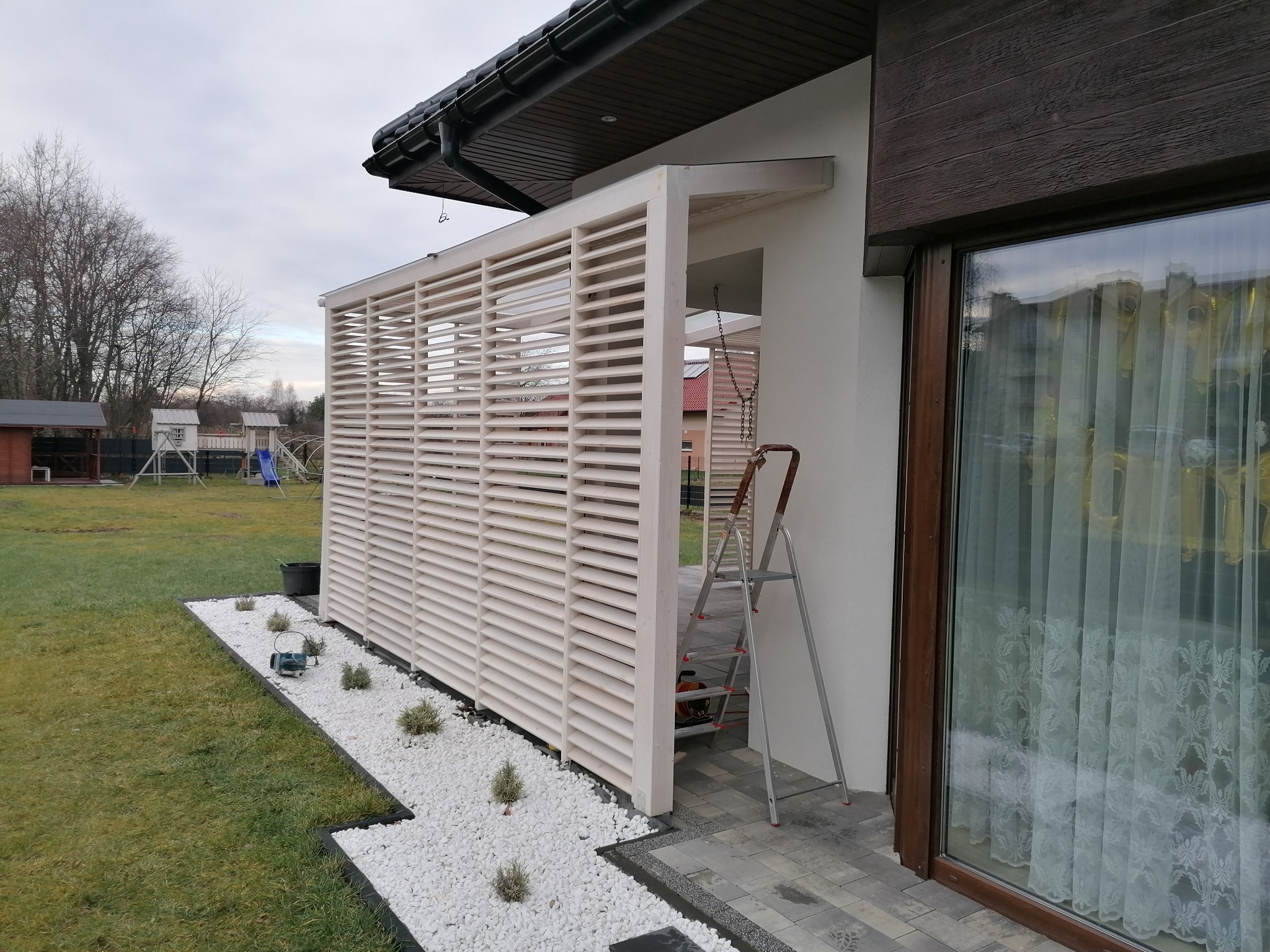 Biała pergola z poziomymi lamelami, częściowo osłaniająca taras, z drabinką opartą o ścianę i dekoracyjnym obłożeniem z białych kamieni i zielonych roślin.