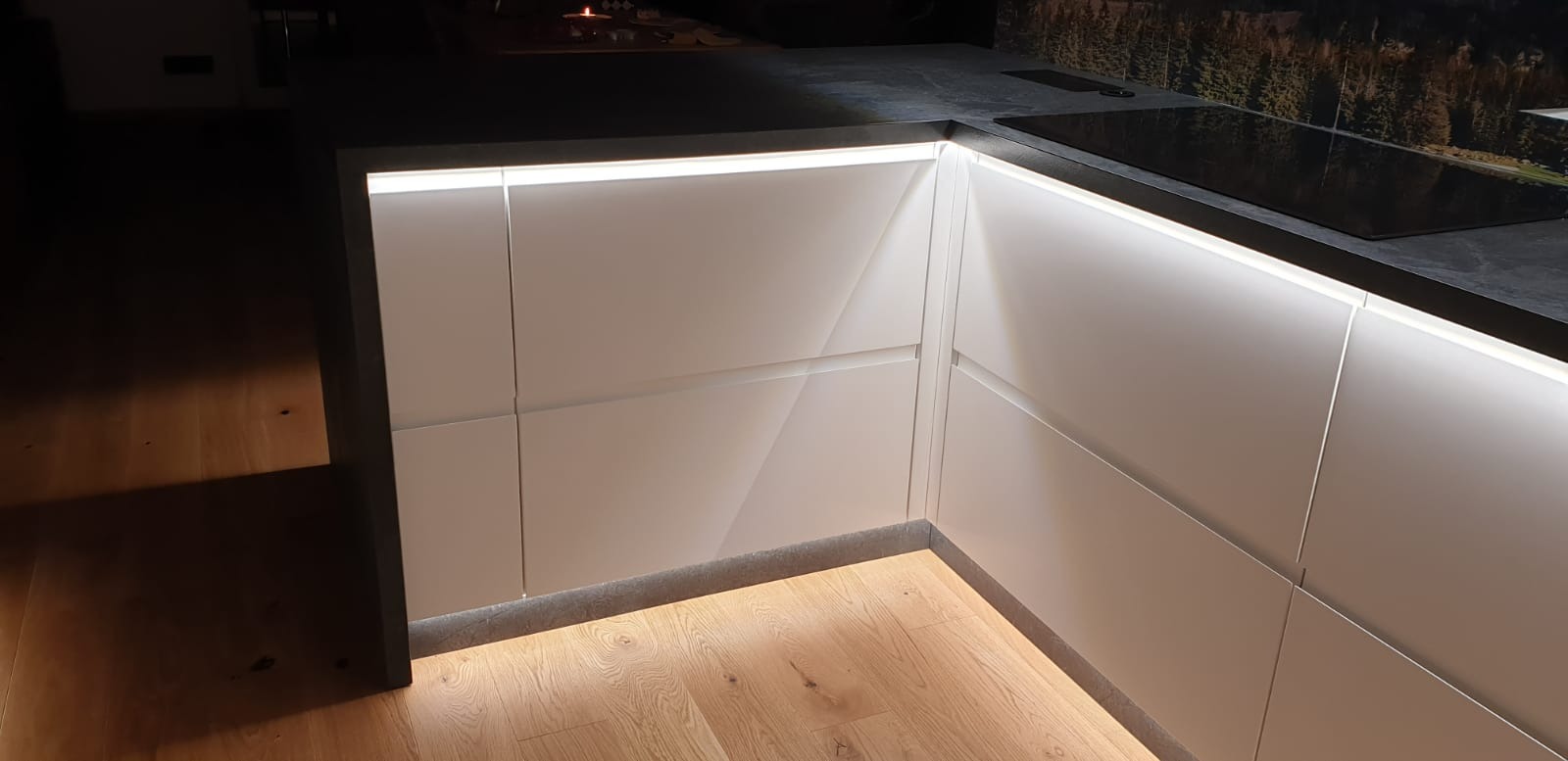 Nowoczesna kuchnia narożna z białymi, matowymi frontami, podświetleniem LED pod blatem i na podłodze, oraz blatem z ciemnego kamienia.