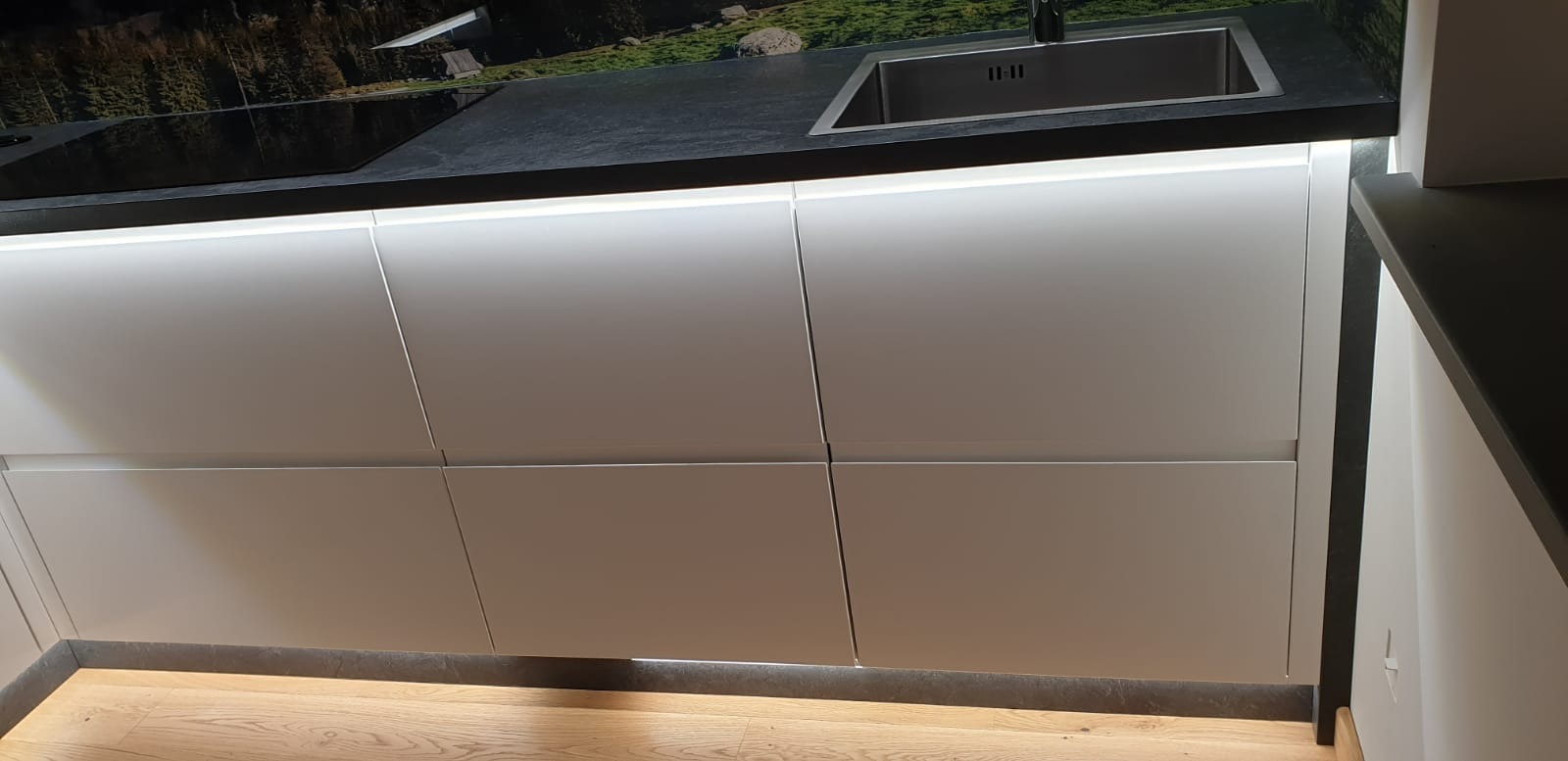 Nowoczesna kuchnia z białymi, gładkimi frontami szafek, podświetleniem LED i blatem imitującym kamień z wbudowanym zlewem i płytą indukcyjną. Na blacie widoczna grafika przedstawiająca górski...