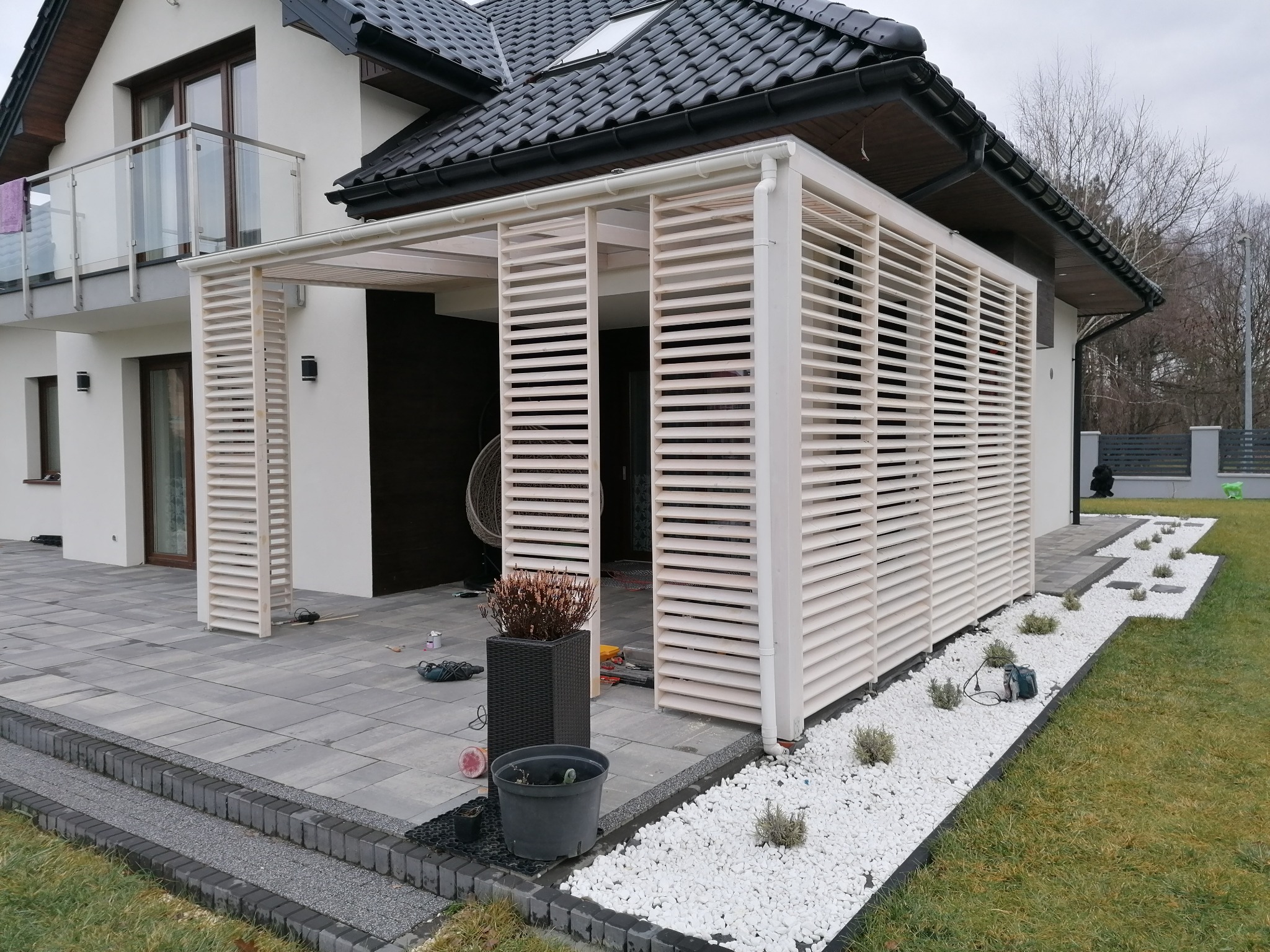 Nowoczesna pergola z białymi lamelami, przylegająca do białego domu z ciemnym dachem, otoczona szarą kostką brukową i białym żwirem z nasadzeniami, widoczne narzędzia i donice.