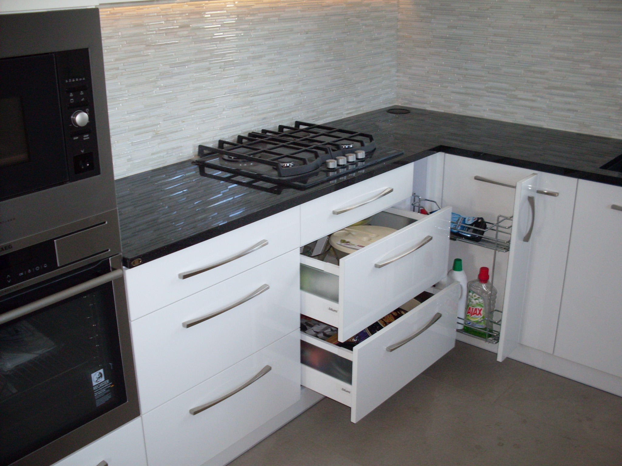 Nowoczesna biała kuchnia z czarnym blatem, otwarte szuflady ukazują zawartość, w tle mozaika na ścianie i płyta grzewcza.