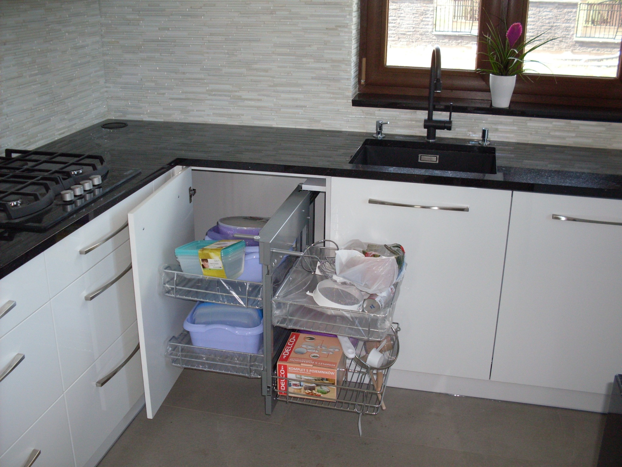 Nowoczesna kuchnia z białymi frontami, czarnym blatem i płytkami imitującymi cegłę. Widoczny otwarty szafka z metalowymi koszami wysuwanymi, zawierającymi różnorodne pojemniki i akcesoria kuchenne.